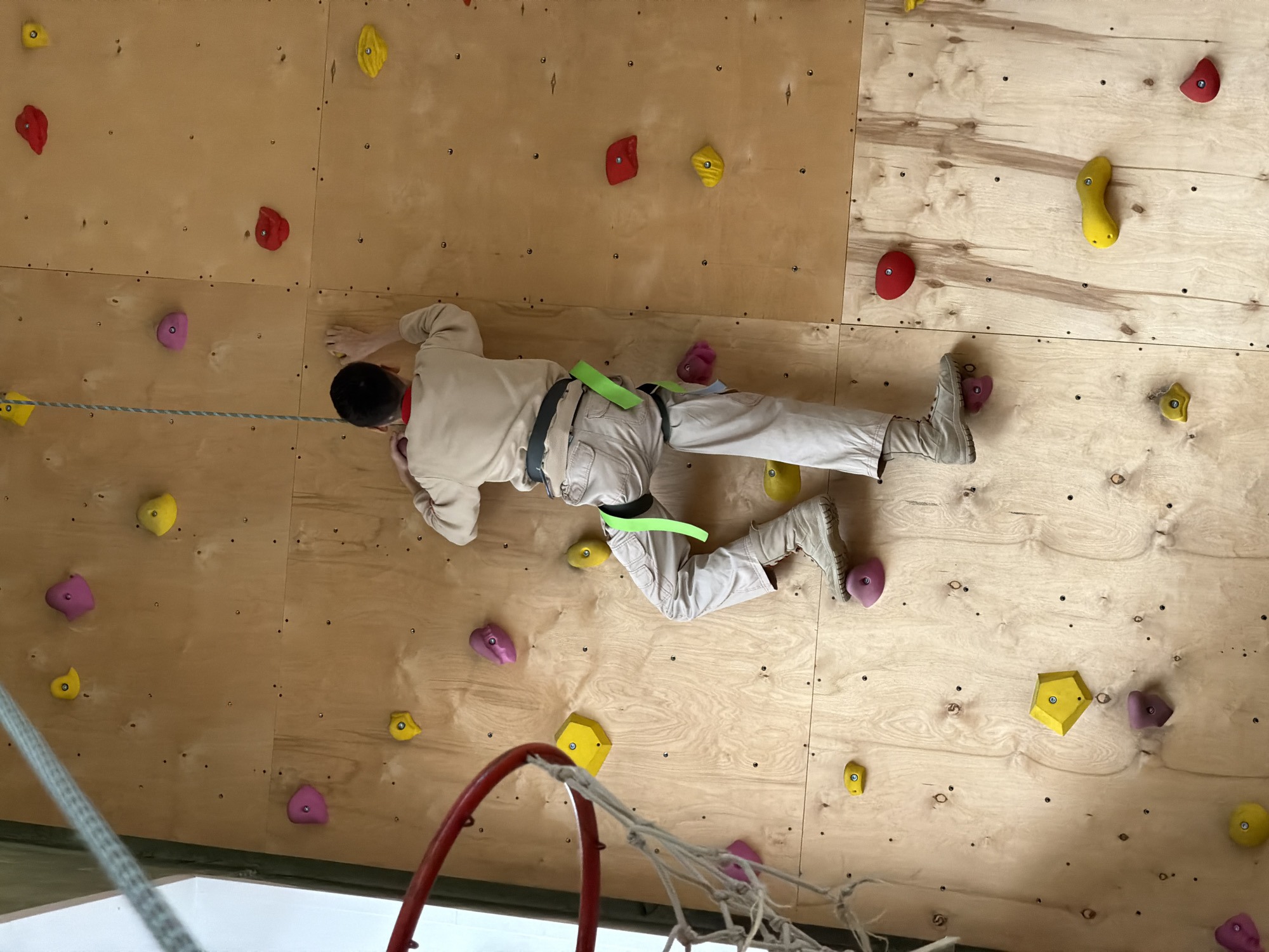 A person is climbing an indoor rock climbing wall. The person is wearing light colored clothing, boots, and a safety harness. The climbing wall is made of wood and has climbing holds of various colors (red, yellow, pink). A rope is visible attached to the person's harness and secured to the wall. A net is at the bottom. The location is Izhevsk, Russia.