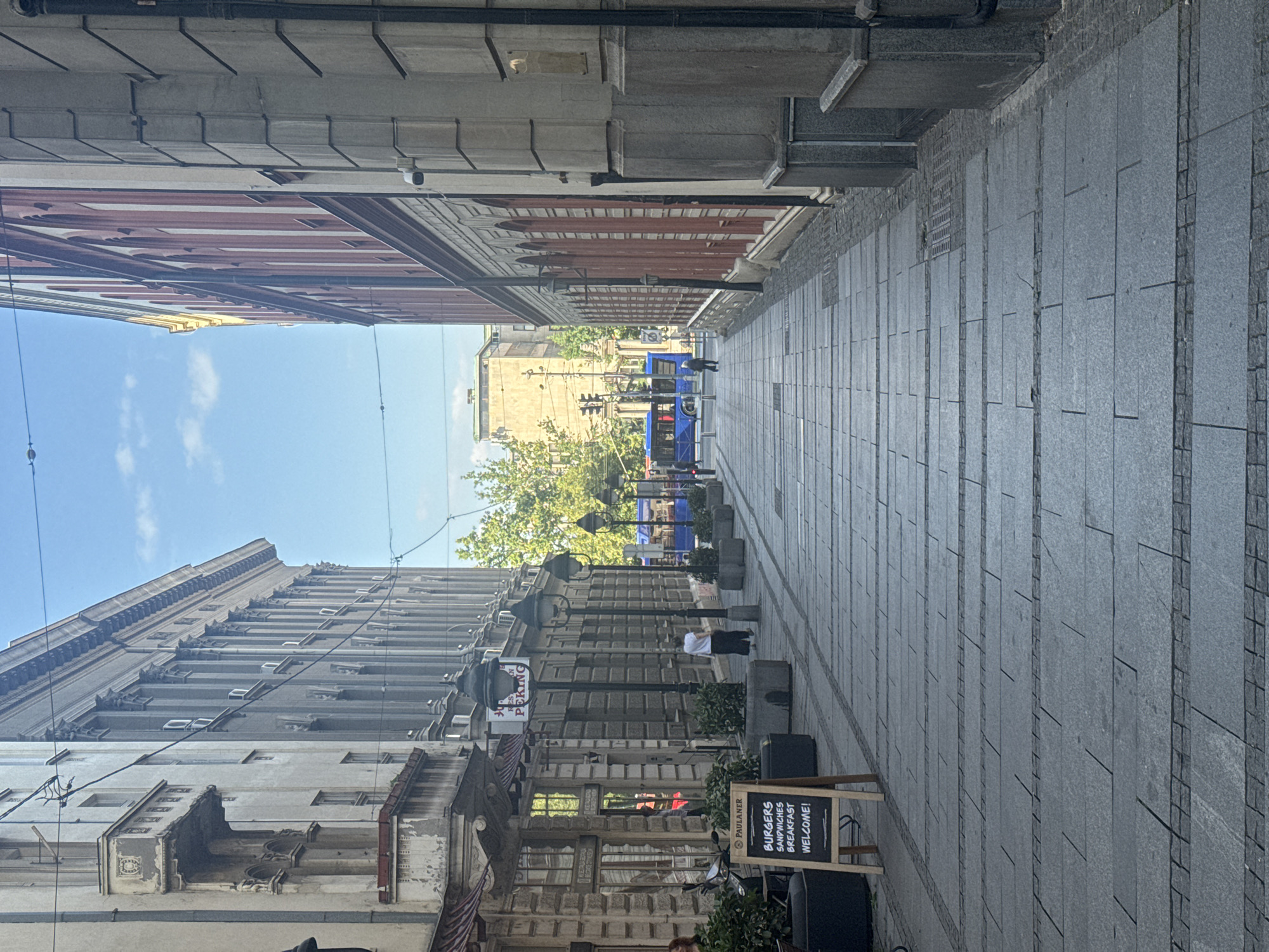 The image depicts a street scene in Belgrade, Serbia, oriented vertically. The perspective looks down a cobblestone pedestrian walkway lined with buildings on either side. The architecture suggests an urban European setting, with multi-story buildings featuring classical or neo-classical facades.
On the left side of the street, a building with a light-colored facade is prominently visible, showcasing numerous windows and decorative architectural elements. Further down the street, on the right, is a similar building with a gray stone facade. The walkway itself is paved with large, rectangular gray stones.
Several objects are scattered along the walkway, including decorative planters with greenery, street lamps, and a black A-frame chalkboard sign. The chalkboard displays white text that reads "BURGERS," "SANDWICHES," "BREAKFAST," and "WELCOME!".
In the distance, a blue trolleybus is visible, indicating public transportation is in operation. A few people are present on the street, though their features are not distinct due to the distance and image quality. One person in a white shirt stands near the trolleybus, and another is seen further down the walkway.
The sky above is blue with scattered white clouds, suggesting a pleasant daytime weather. The overall atmosphere is calm and ordinary, depicting a typical day on a busy pedestrian street in a city. The presence of the trolleybus and the restaurant sign suggests this is an active commercial area.