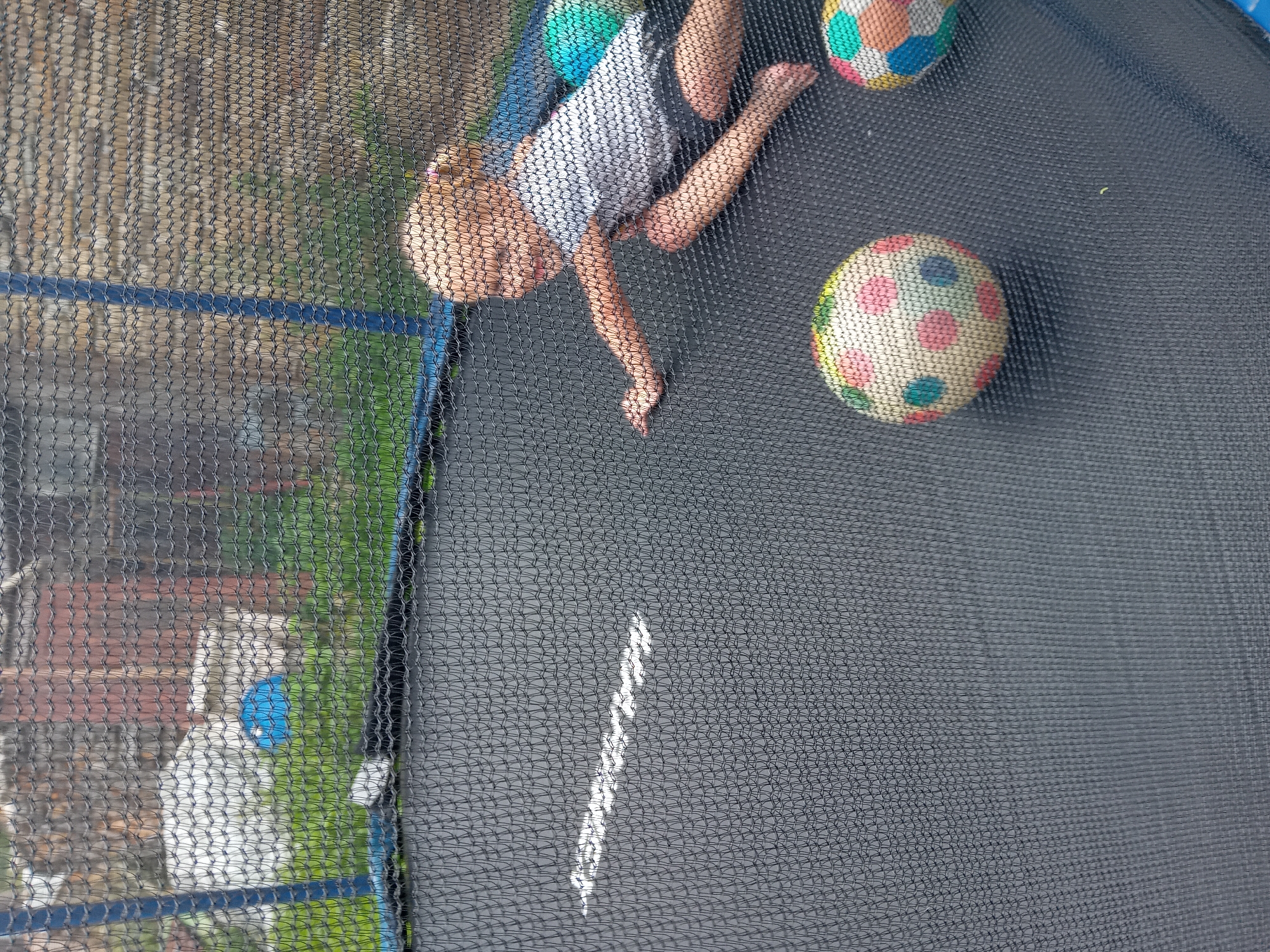 A young, blonde-haired girl is lying on her stomach on a trampoline, reaching out towards the left side of the frame. She is wearing a white t-shirt and dark shorts. Two multi-colored balls are visible on the trampoline, one in the foreground and another further back on the right. The trampoline is enclosed by a black safety net, which is partially obscuring the view of the background. The background appears to be an outdoor environment with green grass and some structures, possibly a fence or wall. The scene is set in Zuberec, Slovakia.