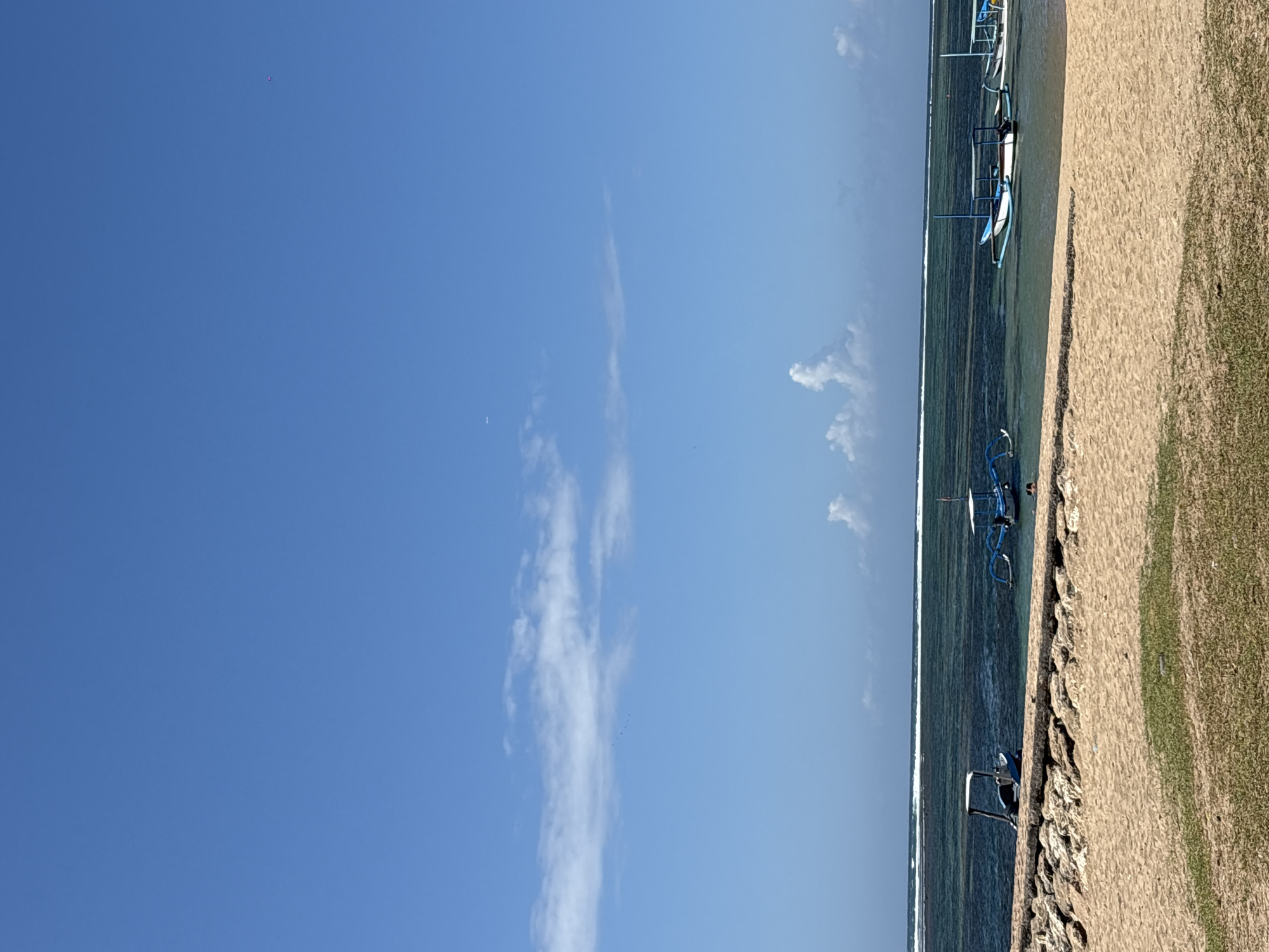 This image depicts a tranquil beach scene at Sanur Beach, Denpasar, Indonesia, under a clear blue sky with scattered white clouds. In the foreground, a sandy beach transitions to a patch of green grass, followed by a breakwater constructed from large, irregular stones.
The shallow, clear turquoise water of the ocean stretches beyond the breakwater. Several traditional Balinese outrigger boats, known as jukung, are visible in the water. Two of these boats are blue and white, with canopies, and appear to be moored or anchored. Another smaller blue boat with a canopy is closer to the shore.
The horizon is marked by a distant line of white surf breaking over a reef. The overall atmosphere is serene and suggests a peaceful day at the beach. There are no prominent human figures, though a distant person may be visible near one of the boats on the right.