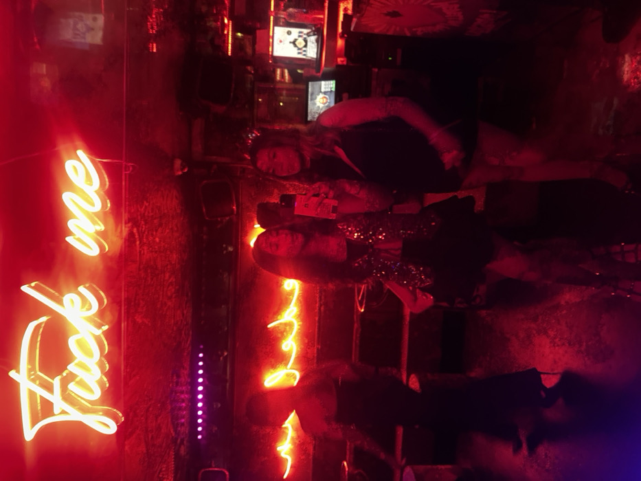 This image depicts the interior of Le Carmen, a nightclub located at 34 Rue Duperré, 75009 Paris, France, in the Pigalle district. The scene is dominated by a pervasive red neon light emanating from various sources.
In the upper center, a prominent yellow-orange neon sign spells out "Fuck me" in a cursive script. Below this, additional linear red neon lights are visible, including what appears to be illuminated Arabic script on a wall or surface. The environment is dark, with reflective surfaces on the floor mirroring the intense red light. Overhead, a security camera is visible.
Three individuals are present. In the mid-ground, two women are positioned. The woman on the left, with long dark hair, holds a smartphone vertically, seemingly engaged in taking a selfie or video. She is wearing a dark top adorned with reflective or sequined material. To her right, a second woman with long hair stands beside her, looking towards the phone. This woman has visible tattoos covering her left arm and is dressed in a dark, possibly sleeveless, garment. In the left background, a third, blurred figure is captured in motion, walking away from the camera, wearing a long dark dress or skirt and high-heeled shoes. On the right side of the background, elements of a bar or DJ booth are visible, including multiple digital monitors, one displaying a black and white checkered pattern and another with green visual elements.