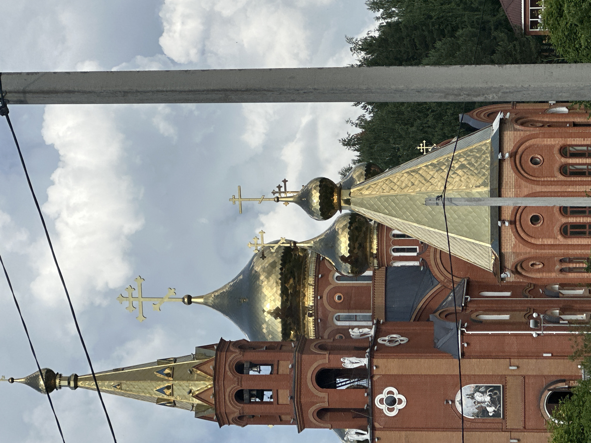 The image shows a close-up view of a red brick church with golden domes and crosses, set against a cloudy sky. The church architecture features multiple domes of varying sizes and shapes, some topped with distinctive Orthodox crosses. A tall, slender concrete utility pole stands in the foreground on the right side, partially obscuring the church. Power lines are visible running horizontally across the upper portion of the frame. The church has intricate architectural details, including arched windows and decorative elements. In the lower left corner, a small mural depicting a figure on horseback is visible on the brick facade. Green trees form a dense background behind the church. The overall impression is of a religious structure within a natural setting, with man-made infrastructure also present. This is the Church of the Intercession of the Holy Virgin in Chaikovsky, Russia.