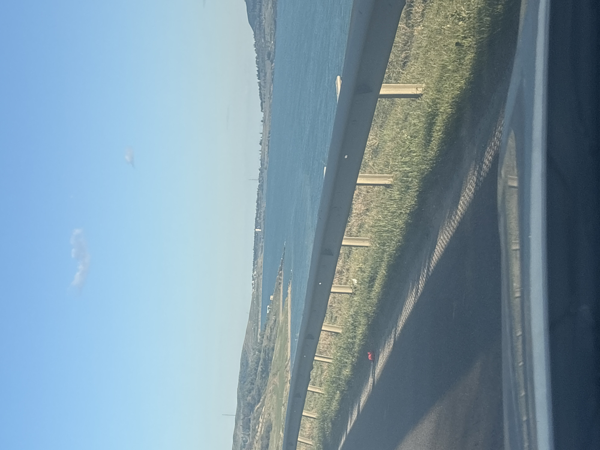 A view from a moving vehicle, likely a car, overlooking a body of water beside a grassy embankment and a roadside barrier. The sky is clear with some wispy clouds. The vehicle's window frame is visible. The location cannot be determined to be Tbilisi, Georgia, based on the visual information available.