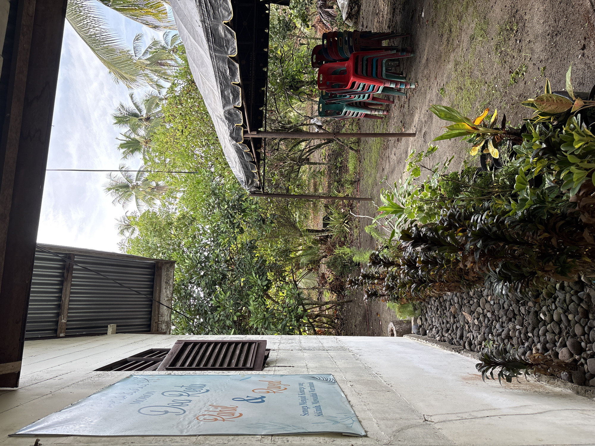 The image depicts an outdoor scene, likely in a rural or semi-rural setting, with elements suggesting a gathering or event. On the left, a white concrete wall is visible, with a blue banner displaying text in a script font, partially obscured by a louvered window. The banner appears to be decorative, possibly for a wedding or celebration, with names like "Rabil" and "Rawal" hinted at. To the right of the wall, a pathway lined with dark, smooth stones leads towards a dense area of various plants and foliage.
In the background, a dark tarped tent is erected, supported by metal poles. Beneath the tent, a stack of colorful plastic chairs is arranged, primarily in red and green, indicating seating for an event. Beyond the tent and chairs, a lush green environment with numerous trees, including palm trees, fills the background, suggesting a tropical or sub-tropical climate. The ground is mostly dirt and dry grass. The sky above is overcast with scattered clouds.
The image does not contain any people or explicit actions. The scene is static and focuses on the setup for an outdoor event and the surrounding natural environment. The location is identified as Beunyot, Indonesia.