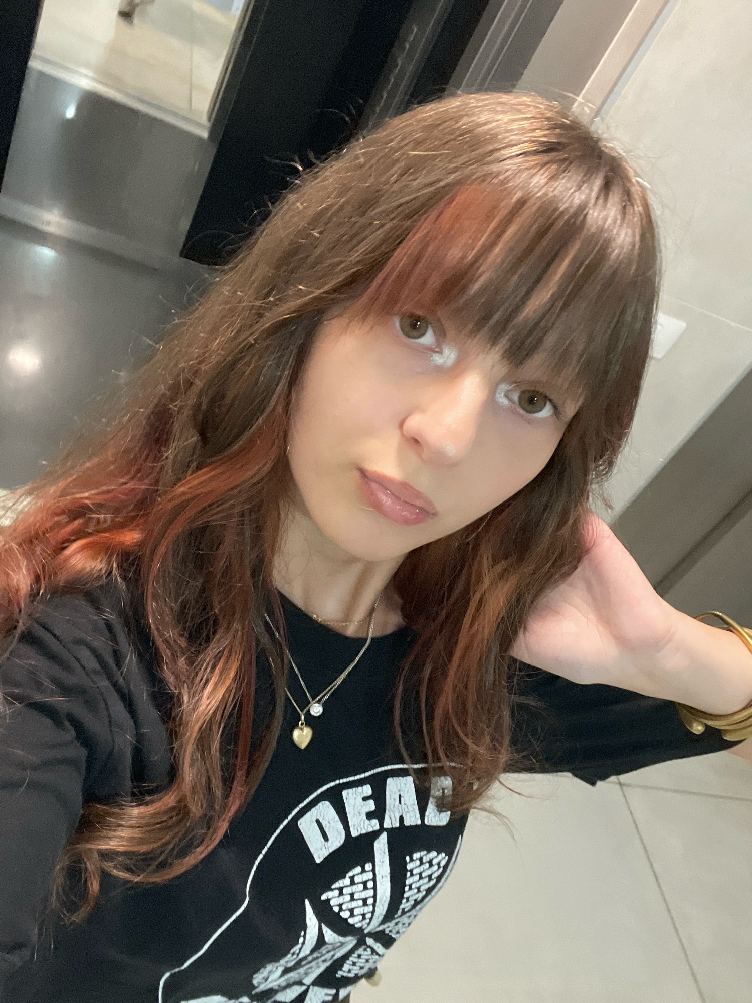 A young woman with long, brown hair, some strands of which are dyed reddish-brown, is taking a selfie in a bathroom. She is wearing a black t-shirt with a white graphic design that appears to read "DEAD" and is accessorized with a gold necklace featuring a heart pendant and a diamond, as well as a thick gold bracelet on her right arm. Her makeup includes noticeable white eyeliner or eyeshadow above her eyes. The background is blurred but suggests a modern bathroom with reflective surfaces, possibly a metal sink or countertop and a tiled floor. The lighting is bright, likely from overhead bathroom lights. The woman is positioned in the foreground, looking towards the camera with her head tilted slightly and her right hand resting on the back of her head. Based on the text and context from other images, this scene is likely located in Raanana, Israel.