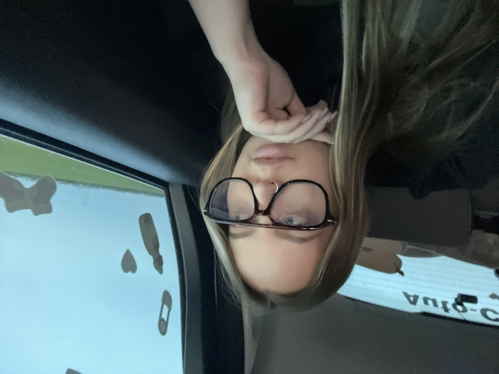 An upside-down close-up photograph captures a young woman inside what appears to be a car. Her face, framed by long, light brown hair, is visible from the forehead to the lips. She wears dark-framed eyeglasses, through which her eyes are visible, and a small nose ring. Her lips are slightly parted. Her right hand, adorned with light-colored, patterned fingernails, rests gently near her forehead.
The background reveals the car's dark interior, likely the headliner and window frame. Through the car window, a bright, light blue sky is visible, with a hint of green along the horizon, suggesting an outdoor, daytime setting. Several white or light-colored decorative shapes are affixed to the car window, including a heart, a baby bottle, and a safety pin. In the lower right corner, a reversed reflection of text, appearing to read "AUTO-C" or similar, is visible on a dark surface, likely a car part or another window. The overall impression is a casual selfie taken in a vehicle during daylight hours.