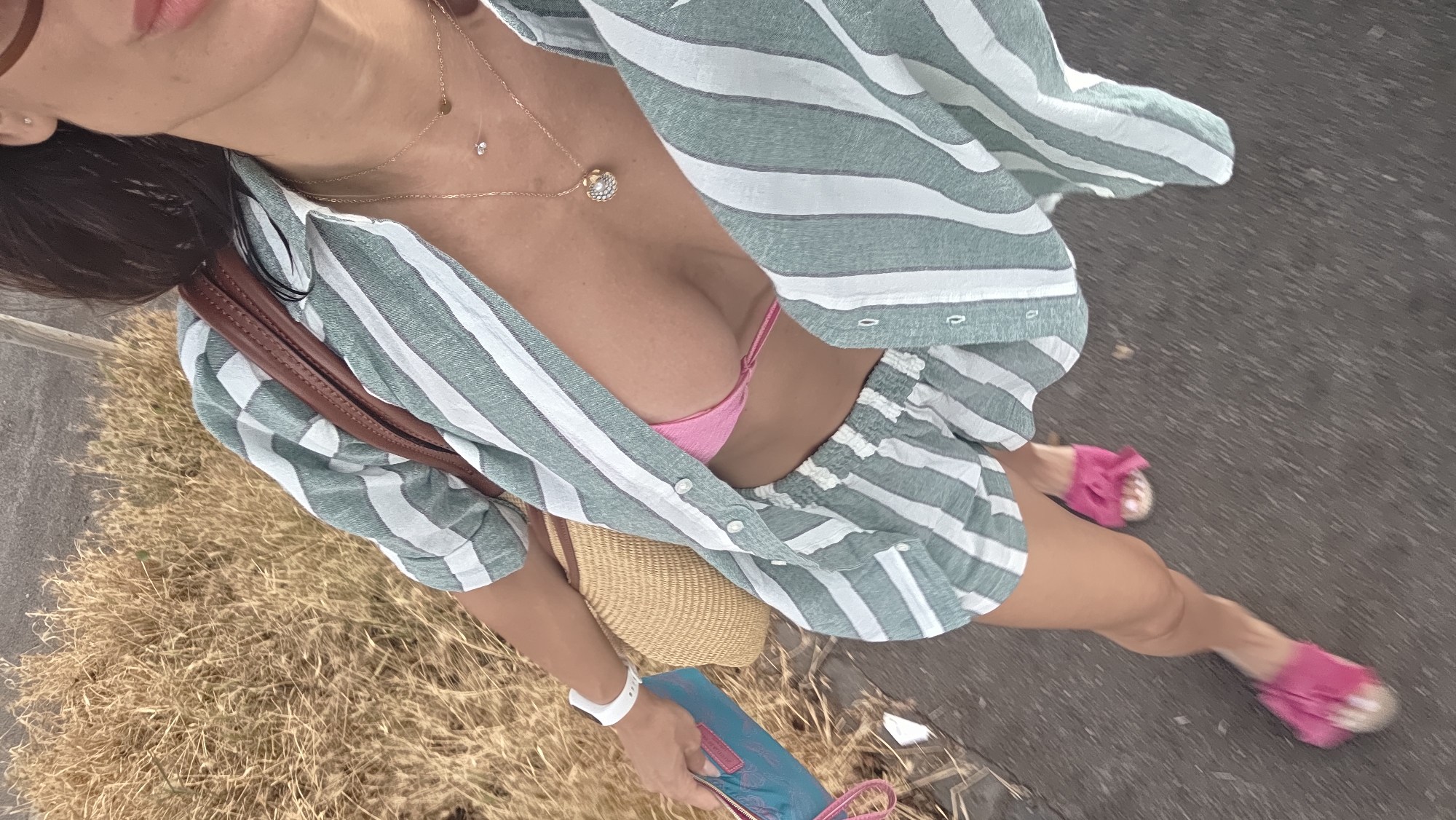 The media file captures a close-up, high-angle, full-body shot of a person walking outdoors. The individual is wearing a green and white striped outfit consisting of an unbuttoned shirt over a pink bikini top, and matching shorts. They are also wearing pink slide sandals. A brown leather purse is slung over their left shoulder, and they are carrying a woven straw bag and a blue clutch. The person has dark hair and is wearing necklaces. The ground appears to be a paved surface next to a patch of dry, straw-like grass. The scene suggests a warm, possibly summery day, with the lighting indicating it could be daytime. The image is taken from a perspective that gives a sense of movement, as if the photographer is walking alongside or slightly behind the subject. There is no visible text in the image. The setting could be a park, a pathway, or a street. The overall impression is one of casual summer fashion and outdoor activity.