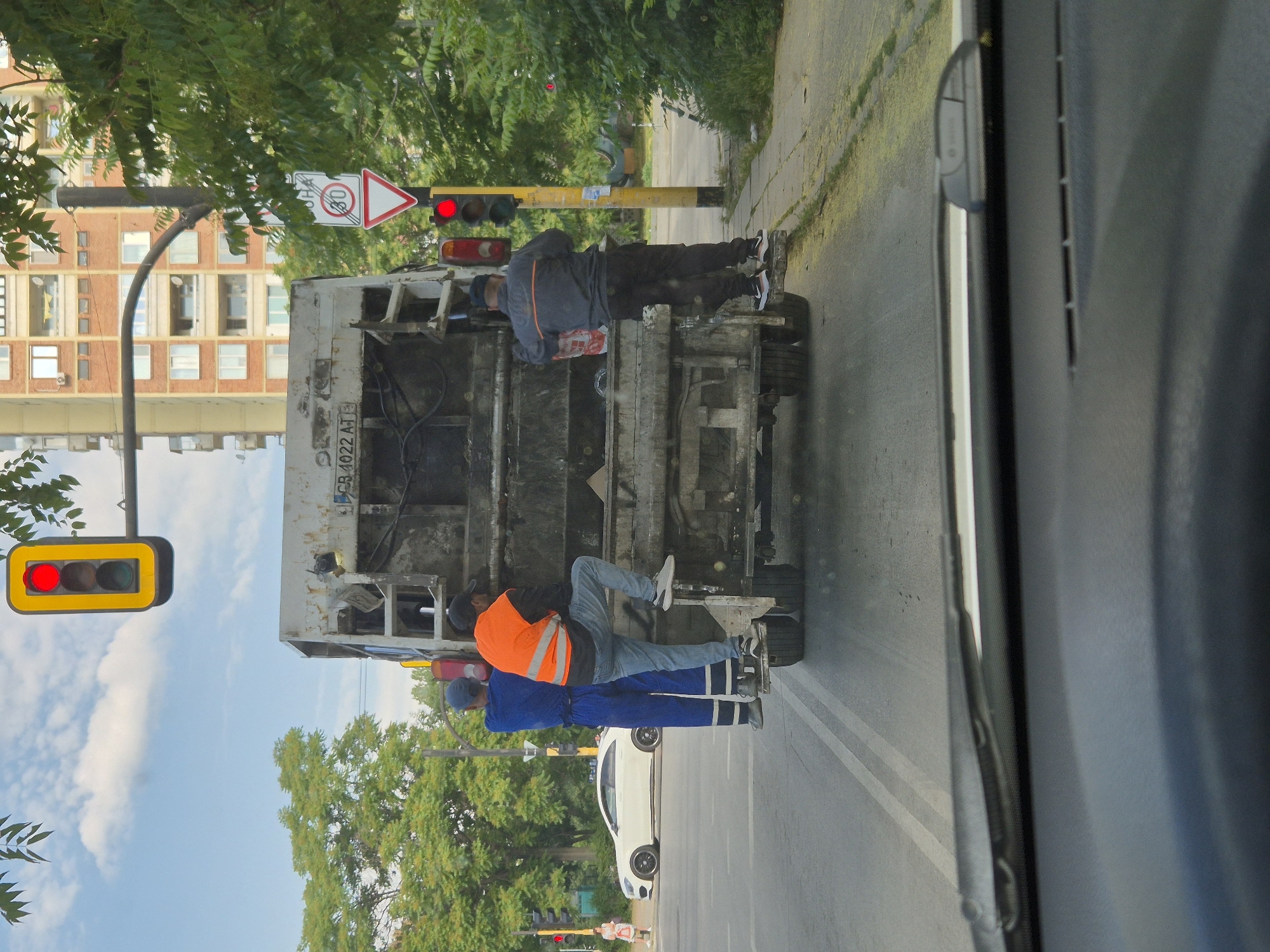 In the city of Sofia, Bulgaria, on a paved street with visible lane markings, a garbage truck is stopped at a traffic light. Three sanitation workers are visible near the rear of the truck. One worker, wearing a blue uniform with white stripes on the pants and a blue cap, stands on the back of the truck, holding onto the frame. Another worker, in a bright orange safety vest and blue jeans, is climbing onto the back of the truck. The third worker, wearing a dark jacket and dark pants, is also on the back of the truck, facing away from the camera, holding a red and white plastic bag. The garbage truck is old and appears worn. In the background, a multi-story brick apartment building stands tall, with balconies on its facade. Green trees line the sidewalk, and a traffic light with a red illuminated signal hangs overhead, along with a circular speed limit sign indicating "30". A white sedan is parked on the side of the road further down. The image is taken from inside a vehicle, with the windshield and dashboard partially visible in the foreground. The sky is partly cloudy.