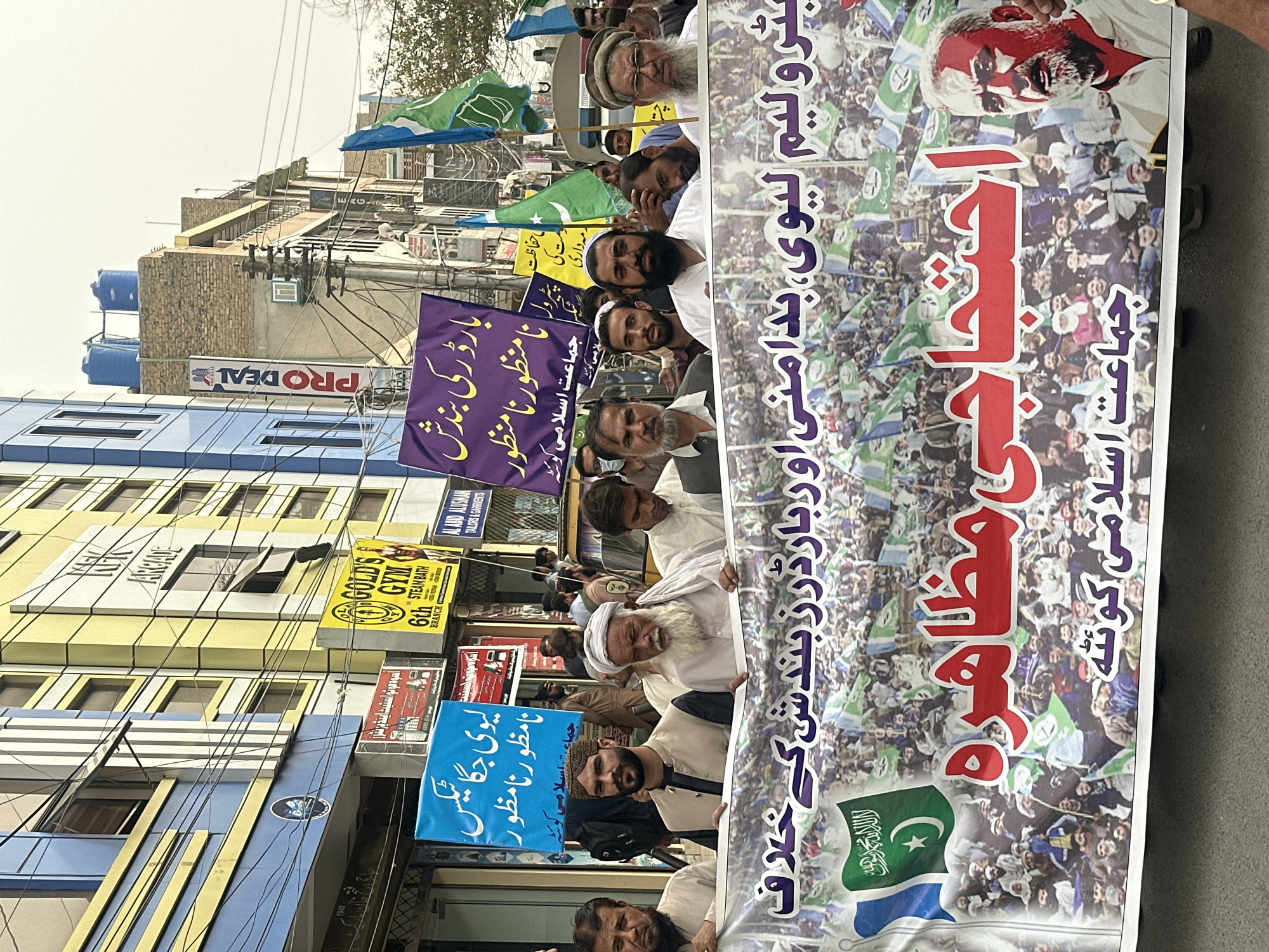 This is a photograph taken in Quetta City Tehsil, Pakistan, showing a street protest. A group of men are holding up a large banner. The banner has writing in Urdu script, some of which reads "بیتولیم بیوی، بدامنی اور درندگی کے خلاف" which translates to "Against Betulim Biwi, Insecurity and Brutality" and "احتجاجی مظاہره" which translates to "Protest Demonstration." Below this, it also says "جماعت اسلامی کوٹه" meaning "Jamaat-e-Islami Quetta."
The people in the foreground are mostly men, with a variety of ages and appearances, some wearing traditional clothing and headwear. The men are holding the banner and looking in different directions, some towards the camera. In the background, there are buildings with shops and signs, some of which are also in Urdu script. One sign reads "GOLD'S GYM." There are also flags visible, including Pakistani flags and green flags with white Arabic script. The street appears to be paved, and there are power lines and utility poles visible. The overall atmosphere suggests a political or social demonstration.