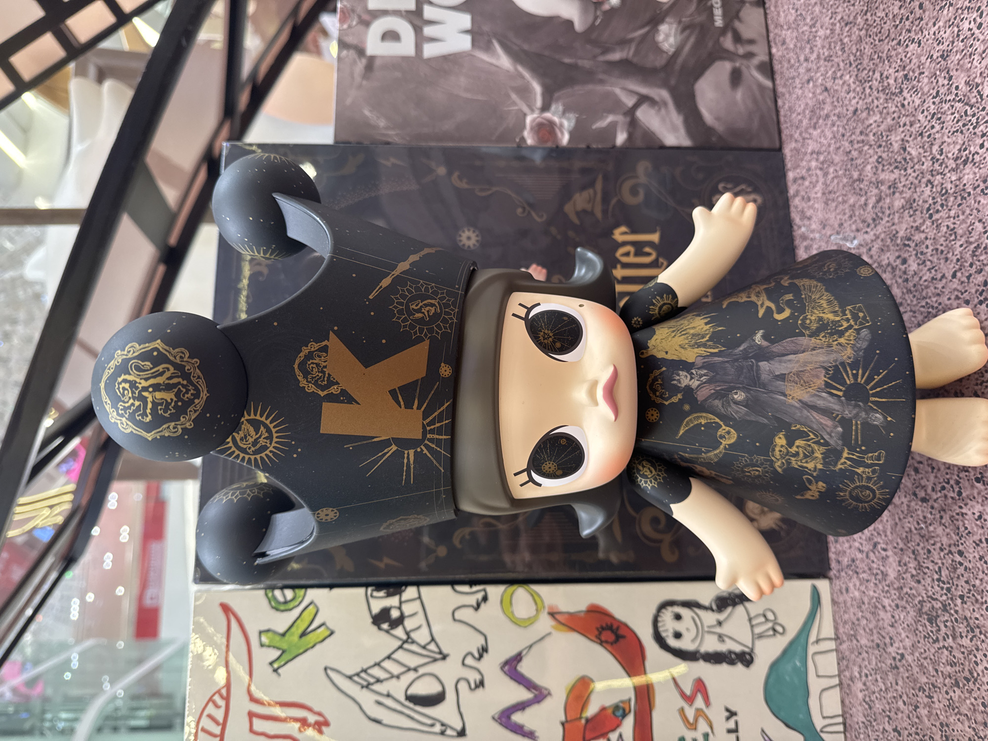 A close-up, eye-level shot features a doll dressed in a black dress with gold celestial patterns and a regal black crown adorned with gold emblems. The doll has large, expressive eyes with intricate golden designs within them, a small pink nose, and soft pink lips. The doll stands in the foreground, with its arms outstretched. Behind the doll, to the left, is a white box with black and green drawings, including the letter "K" and a dinosaur. To the right of the doll are dark boxes with gold patterns, one of which has the partial text "DI W", and another box with a black and gold image of a rose and a figure. The background is blurred, showing reflections of lights and what appears to be shelving. The overall scene is well-lit and appears to be set in a retail or display environment, possibly a toy store or gallery. The specific location in Hanoi, Vietnam, cannot be determined from the visual information provided in the image.