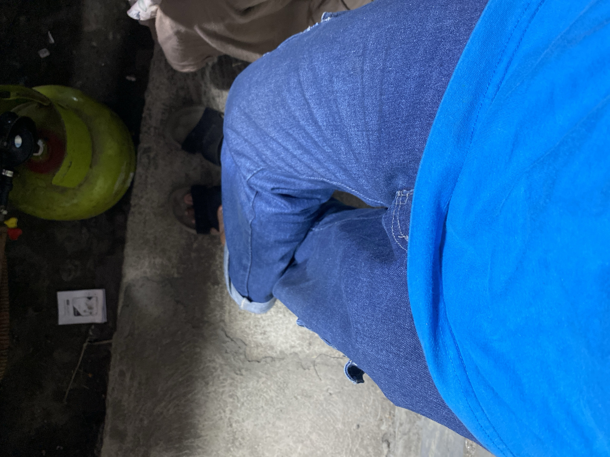 The image shows a close-up, overhead view of a person's legs, clad in blue jeans and a bright blue t-shirt. The person is sitting on a rough, concrete surface, possibly an outdoor area. In the background, a bright green gas cylinder with a red and black regulator is visible, along with a white pamphlet or booklet and some scattered debris. The scene appears to be in an urban or semi-urban environment. The lighting suggests it is either nighttime or a poorly lit indoor space. The location is identified as Kasihan, Indonesia.