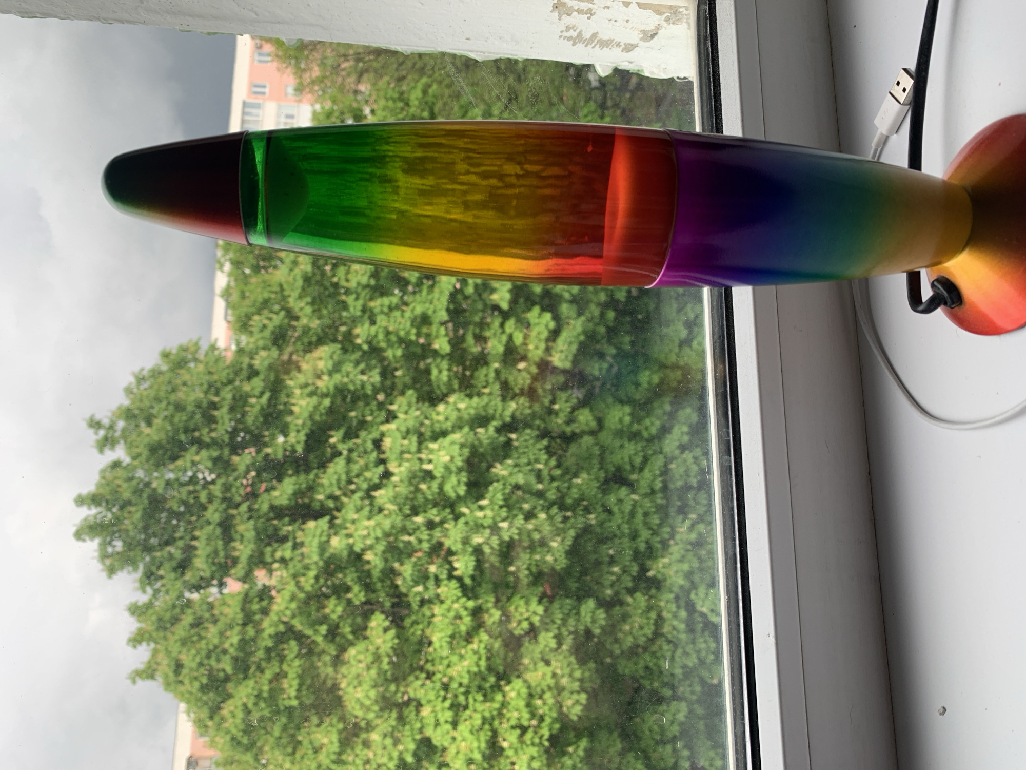 A rainbow-colored rocket lamp lies horizontally on a white windowsill. Outside the window, there is a view of green trees and a cloudy sky. The location is Unknown, Unknown.