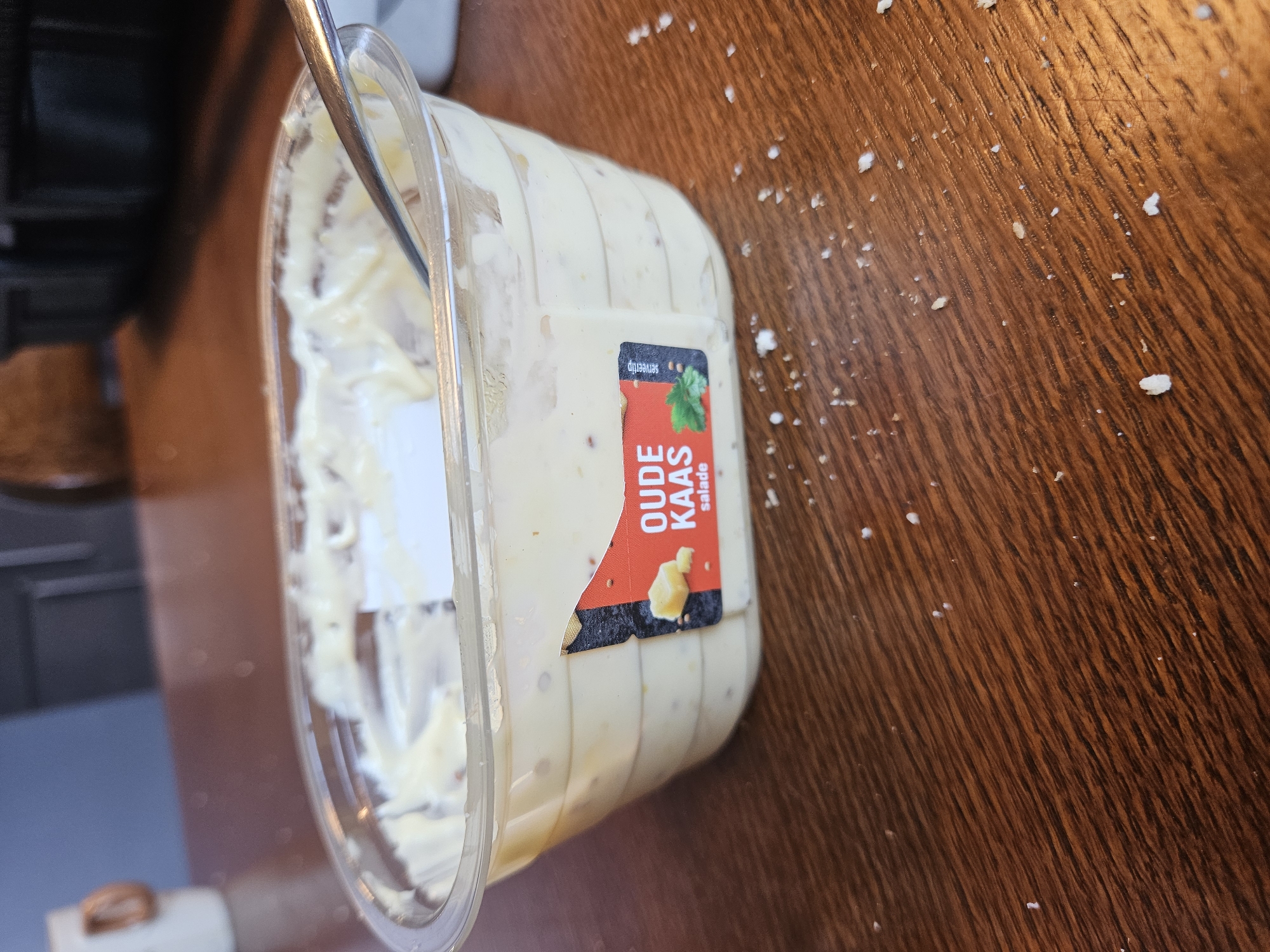 An overhead, slightly angled medium shot captures a clear plastic tub of "Oude Kaas Salade" (Old Cheese Salad) on a brown wooden table. The tub is partially full, with a silver spoon inserted into the creamy yellow salad. A red and black label on the tub displays an image of cheese and herbs, along with text indicating the product's name and type. Scattered across the wooden surface are small white crumbs. The background is blurred, showing hints of kitchen cabinetry and a white mug with a copper handle on a dark surface. The scene appears to be indoors, possibly in a kitchen or dining area in Bemmel, Netherlands.