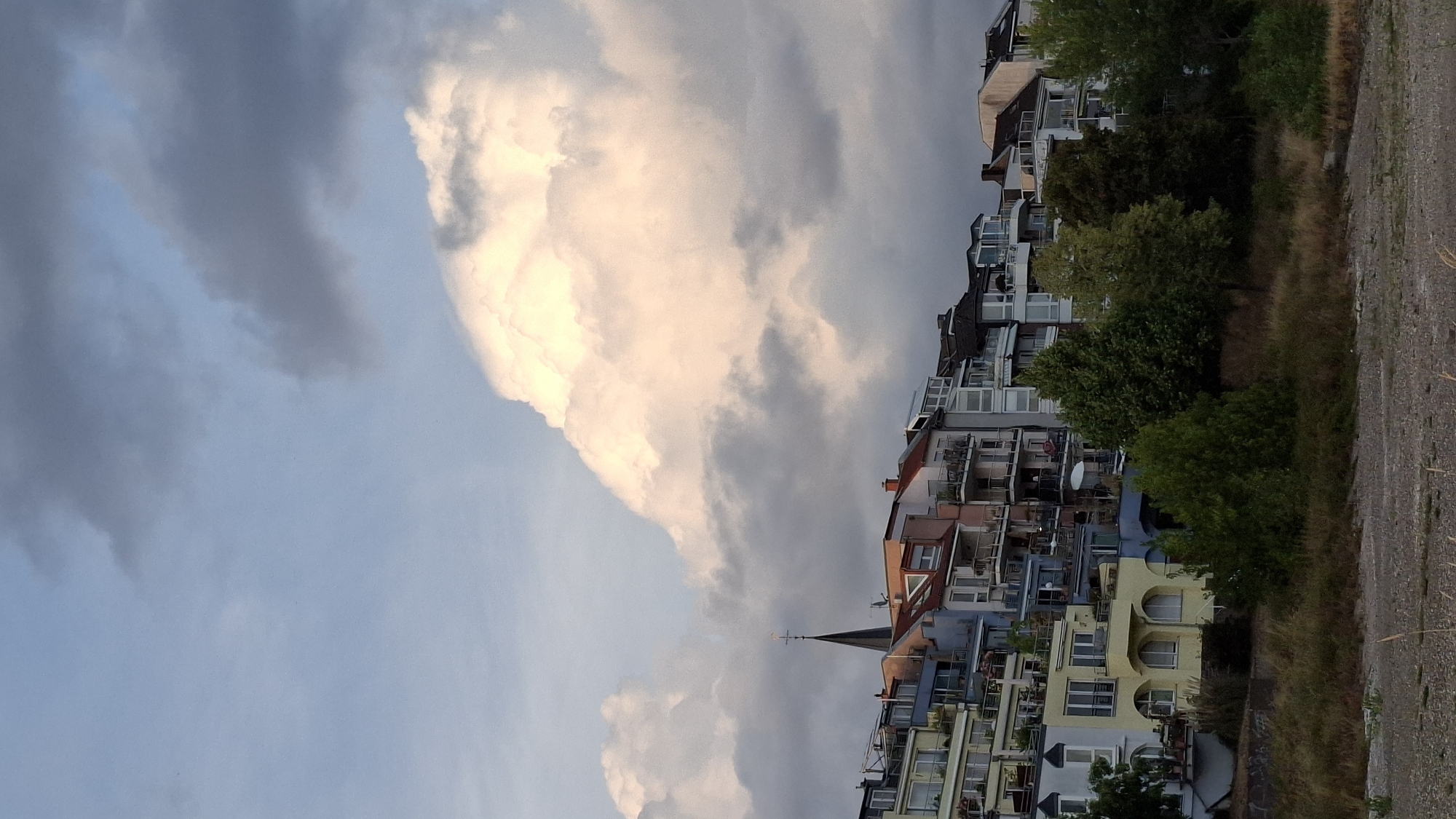 A photograph captured from across the Rhine River in Cologne, Germany, shows a row of residential buildings on the riverbank under a dramatic sky. The buildings are multi-story, with balconies and a variety of architectural styles, suggesting a mix of older and more modern constructions. Prominent among the buildings is a church spire with a cross at its apex. The sky is filled with large, billowing clouds, some brightly lit from behind, while others are darker and more ominous. Trees and vegetation line the riverbank in front of the buildings, and a pebbly shore is visible in the foreground. The lighting suggests it is either early morning or late afternoon.