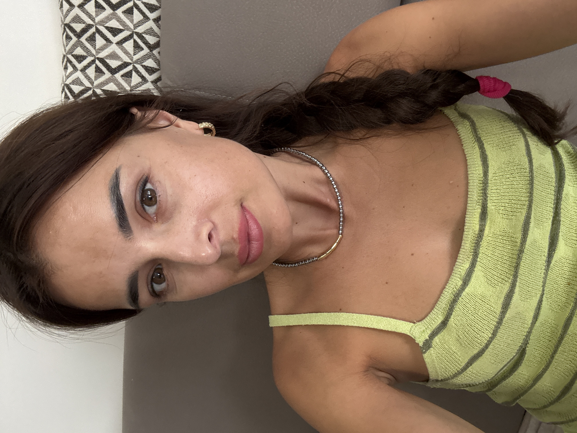 A woman with dark brown hair braided to her right, brown eyes, and light-colored lips is shown in a close-up, forward-facing shot. She is wearing a lime green, sleeveless tank top with horizontal gray stripes and a textured pattern. Around her neck is a silver beaded necklace with a gold accent. She has a gold hoop earring in her right ear. The background is a muted brown, possibly a couch, with a pillow featuring a black and white geometric pattern visible behind her. The lighting is soft.