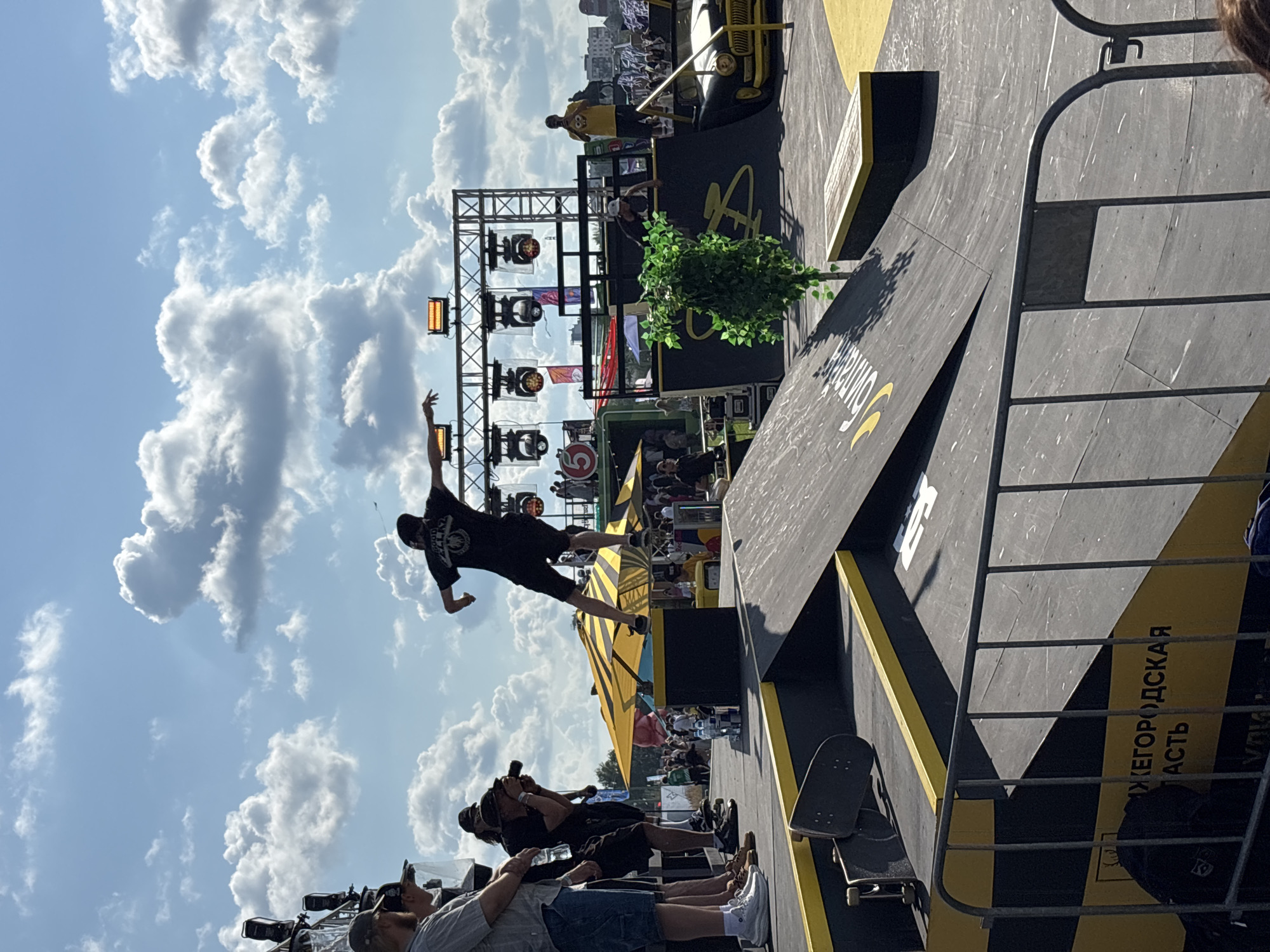 The image depicts a skateboarding event taking place outdoors, likely in the city of Nizhny Novgorod, Russia, as indicated by the visible text "НИЖЕГОРОДСКАЯ ОБЛАСТЬ" (Nizhny Novgorod Oblast) on a black and yellow striped structure in the foreground. The central focus is a skateboarder in mid-air, performing a trick on a ramp. The skateboarder is wearing a black t-shirt and shorts and a black cap. Several spectators are visible on the left side of the image, some with cameras, observing the event. In the background, there is a stage with lighting equipment and a yellow umbrella. A vintage black and yellow car is positioned on the right side of the frame, also part of the event setup. The sky is blue with scattered white clouds.