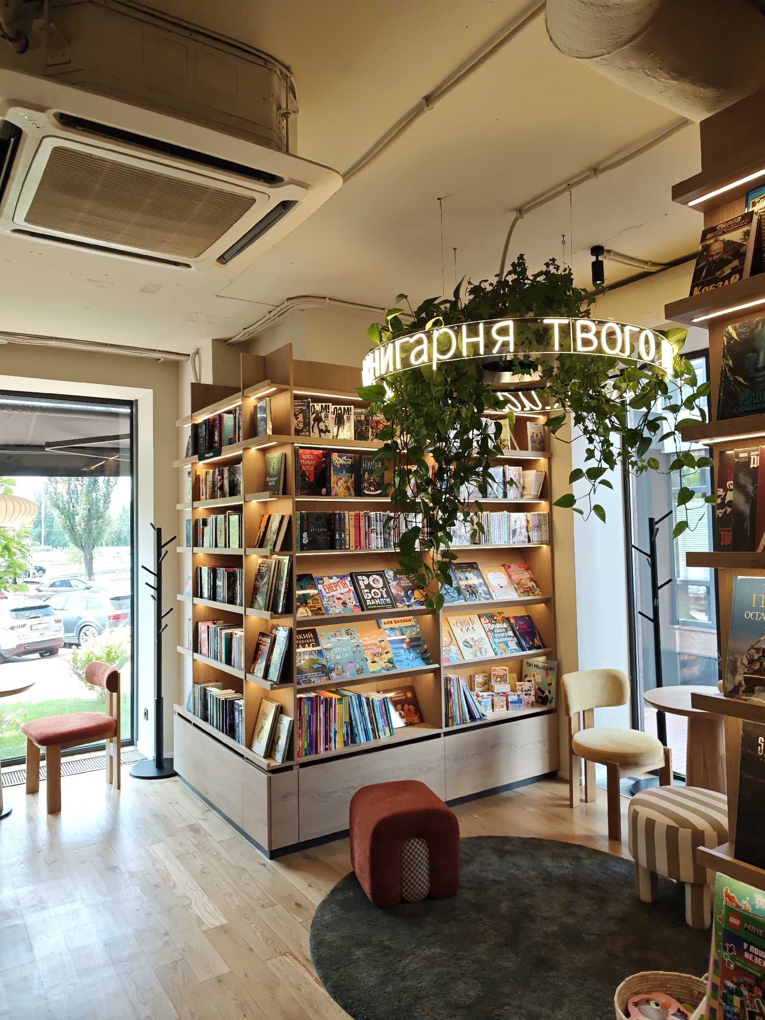 This image depicts the interior of a modern bookstore located in Kyiv, Ukraine. The store features a large, floor-to-ceiling bookshelf filled with various books, some of which are illuminated by overhead lighting. Suspended from the ceiling is a circular neon sign with Cyrillic lettering that reads "Книгарня Твого" (Bookstore of Yours), surrounded by lush green hanging plants. In the foreground, there are several pieces of contemporary furniture, including an orange upholstered stool, a striped pouf, and a yellow upholstered chair. A dark green rug is placed on the light-colored wooden floor. Through a large window on the left side of the frame, a street scene with parked cars and trees is visible. The overall atmosphere is cozy and inviting, suggesting a space designed for reading and relaxation.