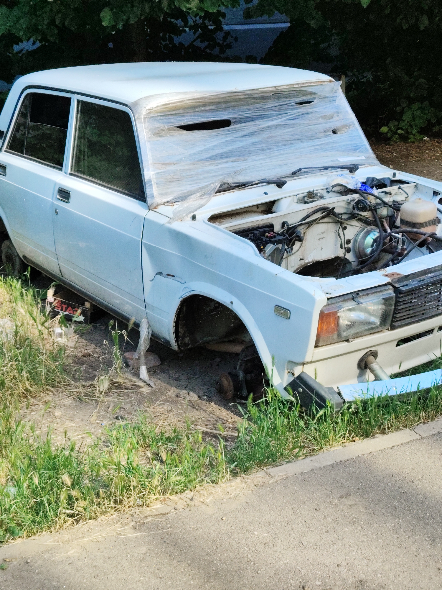 An abandoned white sedan is parked on the side of a paved road in a grassy area, likely in Krasnodar, Russia. The car is in a state of disrepair. Its hood is open, revealing the engine and various wires. The windshield is covered in clear plastic wrap, which appears to have holes. The front passenger-side tire is missing, and the car is supported by what appears to be a wooden block and possibly a jack stand. The car's front bumper is askew. The surrounding environment consists of green grass, dirt, and trees in the background. The lighting suggests it is daytime.