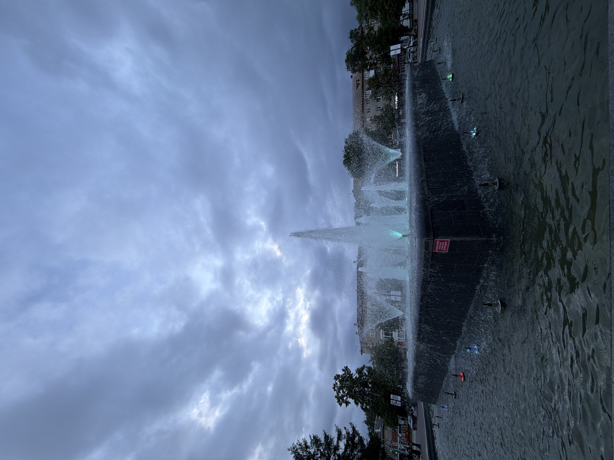 An evening long shot captures a large, illuminated fountain in the city of Chaikovsky, Russia, under a heavily overcast, dull blue-gray sky. The multi-tiered fountain, composed of dark, likely stone, rectangular and angular basins, occupies the lower and central foreground. Its central structure features a tall, narrow jet of water, flanked by multiple smaller, arcing jets. All water streams are brightly illuminated with blue and cyan lights, with some submerged lights in the lower basin emitting blue, green, and red hues. Water cascades over the dark upper basin, creating a shimmering effect. A red and white warning sign with Cyrillic text is affixed to the right side of the fountain's central wall. In the background, an urban environment is visible, with multi-story buildings, dark green deciduous and coniferous trees, and a street. On the left side, indistinct figures of people walk on a sidewalk, and several cars are parked or moving. Various signs with Cyrillic text are visible on buildings and poles in the midground, including "Аптека" (Pharmacy) and "СГУ". The overall scene suggests a public square or park area at dusk or early night, characterized by artificial lighting against a dim natural backdrop.