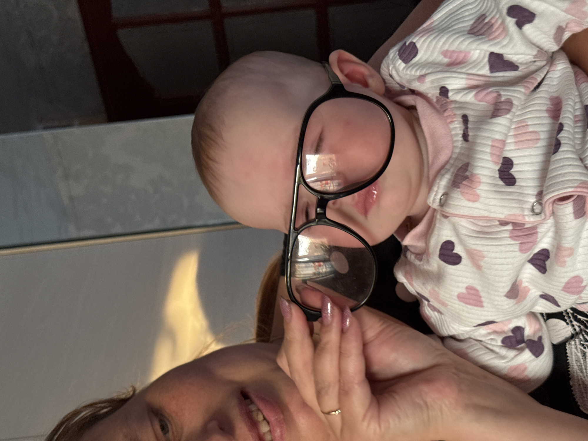 The image shows a close-up of a baby wearing oversized glasses, held by an adult. The baby's face is partially obscured by the large lenses of the glasses. The baby is wearing a white onesie with a pattern of pink and purple hearts. The adult's hand is visible, holding the glasses on the baby's face, and their fingernails are painted with glitter. The background is softly lit, with hints of natural light filtering in, suggesting an indoor setting. The overall impression is a playful and intimate moment between the adult and the child. The image does not contain any specific elements that would allow for the determination of its location in Kemul, Russia.