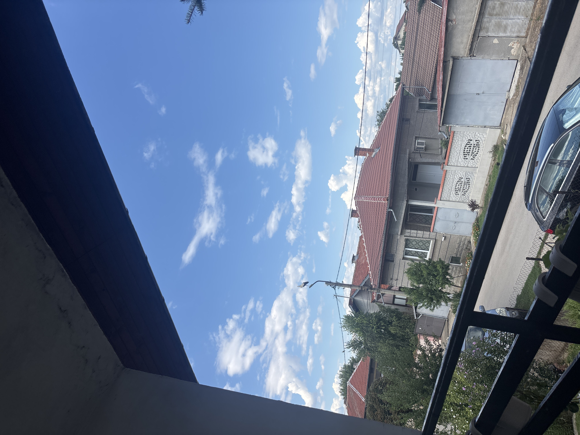 The image captures a view of a residential street in Marten, Bulgaria, from a slightly elevated vantage point, possibly a balcony or window. The sky is a vibrant blue with scattered white cumulus clouds. Dominating the foreground is the underside of a dark overhang, likely a roof or balcony, casting a shadow.
Below the overhang, a neighborhood of houses with red tile roofs is visible. Some houses have light-colored walls, while others appear to be made of concrete. A distinctive feature is a street lamp with a stork perched on top, a common sight in rural and semi-rural Bulgarian areas. The street itself is paved, and several cars are parked along its side, including a dark-colored sedan and a larger, darker vehicle.
Fencing and vegetation, including trees and flowering bushes, line the street and the properties. One house features a decorative white fence with an intricate pattern. Electrical wires are strung across the scene, typical of urban or suburban infrastructure. The overall impression is of a peaceful, ordinary day in a residential area of Marten.
