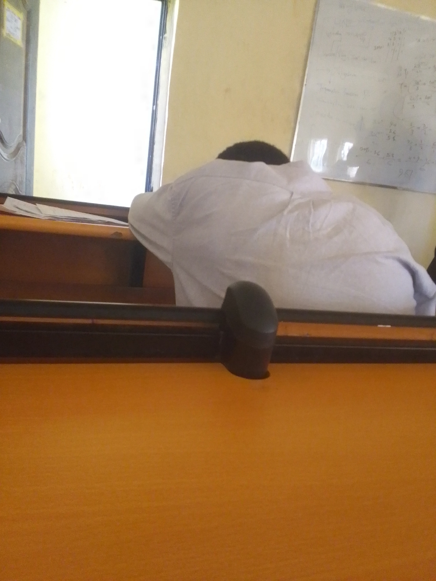 The image shows a person with their head down on a desk in what appears to be a classroom or study hall. The person is wearing a white garment, possibly a shirt or robe. The desk is made of wood and has a dark brown edge. A black computer mouse is visible on the desk in front of the person. In the background, there is a whiteboard with some writing on it, likely mathematical equations or notes. The walls are a pale yellow color. To the left of the image, a partially visible door or partition can be seen, with a bright light source behind it, possibly a window or another room. The overall scene suggests a quiet and perhaps studious environment, although the person's posture might indicate fatigue or boredom. The time of day and weather are not discernible from the image. No specific emotions are clearly visible due to the person's position. There is some faint, illegible text on the whiteboard.