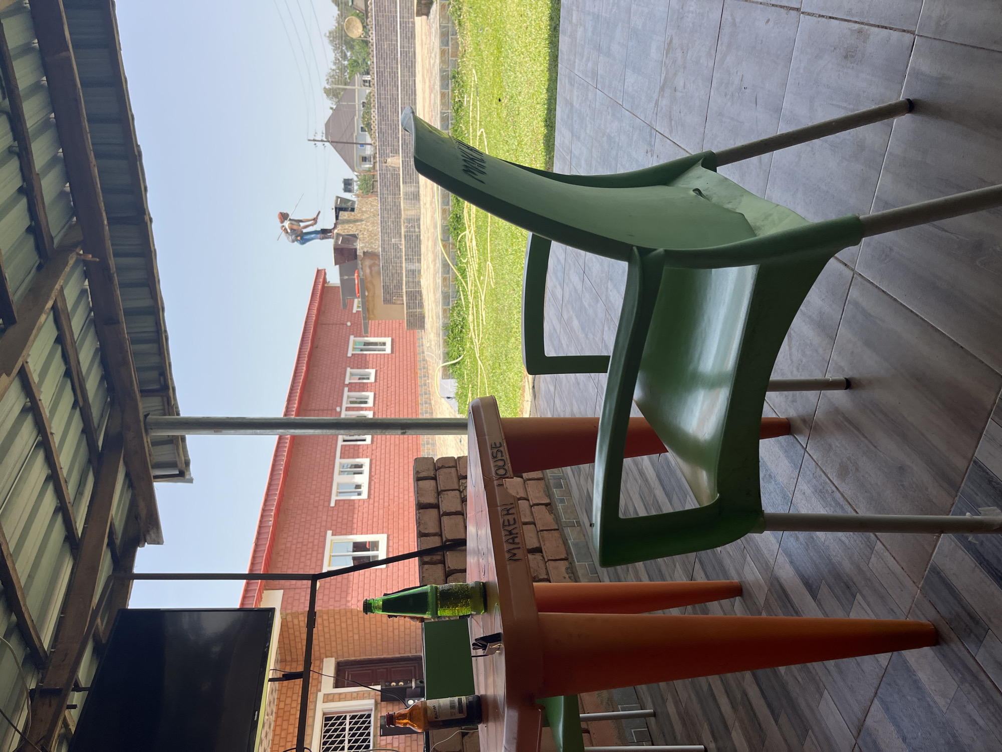 The image depicts an outdoor seating area at "Maker House" in Jos, Nigeria. In the foreground, a green plastic chair is positioned in front of an orange plastic table. The table has the words "MAKER HOUSE" scrawled on its surface. Two bottles, one amber and one green, sit on the table. A television is mounted on a pole to the left. In the background, a red brick building with white-framed windows is visible. Beyond the building, a person is seen working on a construction project. The scene is set on a sunny day with a clear sky.