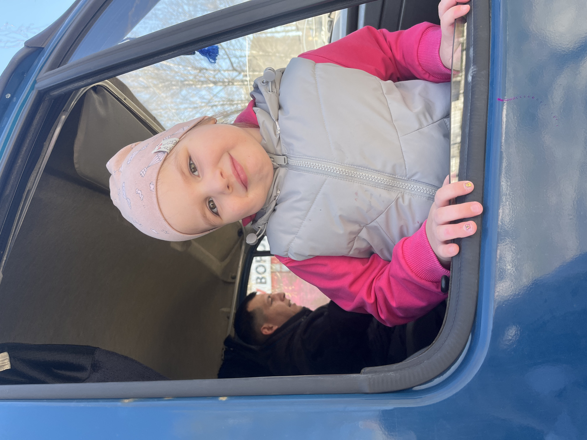 A young girl in a pink hat, pink shirt, and gray vest is peering out of a truck window. A man is resting inside the truck. The window frame and truck's exterior are blue. A sign with Russian text is visible in the background, indicating a location in Izhevsk, Russia.