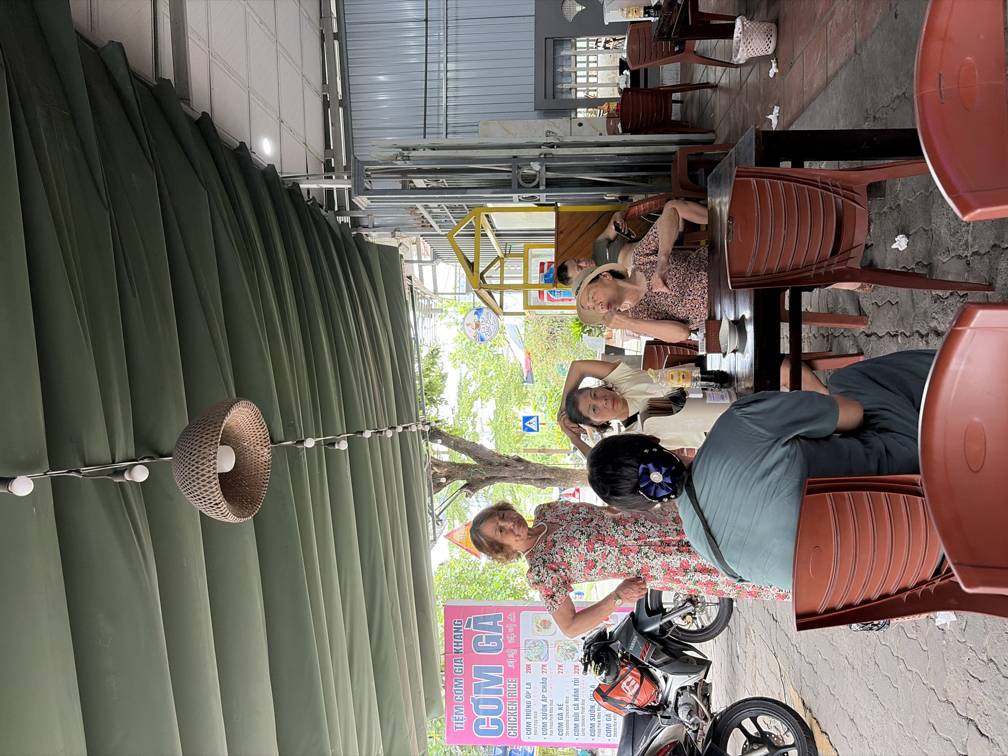 The image captures an outdoor seating area of a Vietnamese restaurant, identified as "Tiệm Cơm Gia Khang" by a prominent pink sign. The setting is likely in the city of Đà Nẵng, Vietnam, given the presence of Vietnamese text and the overall urban outdoor dining aesthetic common in the region.
In the foreground, four individuals are seated around a dark wooden table. The person closest to the viewer is wearing a teal-colored top and dark pants, with their back to the camera. They have a dark blue bow with a flower accent in their hair. To their left, an older woman in a floral print dress with a pearl necklace stands and looks towards the camera, appearing to be addressing the viewer or engaging with someone off-camera. To her right, a woman with short dark hair wears a light-colored t-shirt and rests her hand on her head. Across the table, another woman in a patterned short-sleeved shirt is seated, with her right hand raised to her mouth, appearing to be eating or in conversation.
The environment is partially covered by a large, dark green tarp stretched overhead, providing shade. Several string lights with exposed bulbs hang from the tarp, along with a larger woven lampshade. To the left of the frame, a portion of a black motorcycle with an orange and black backpack is visible. In the background, a light blue and yellow structure, possibly a small kiosk or decorative element, is present. Further back, metal fencing and corrugated metal walls suggest the proximity of other buildings. The ground is paved with interlocking gray tiles. Scattered on the ground are pieces of white paper, likely napkins or food waste. A white woven trash bin is visible to the right. The overall scene depicts a casual, open-air dining experience.