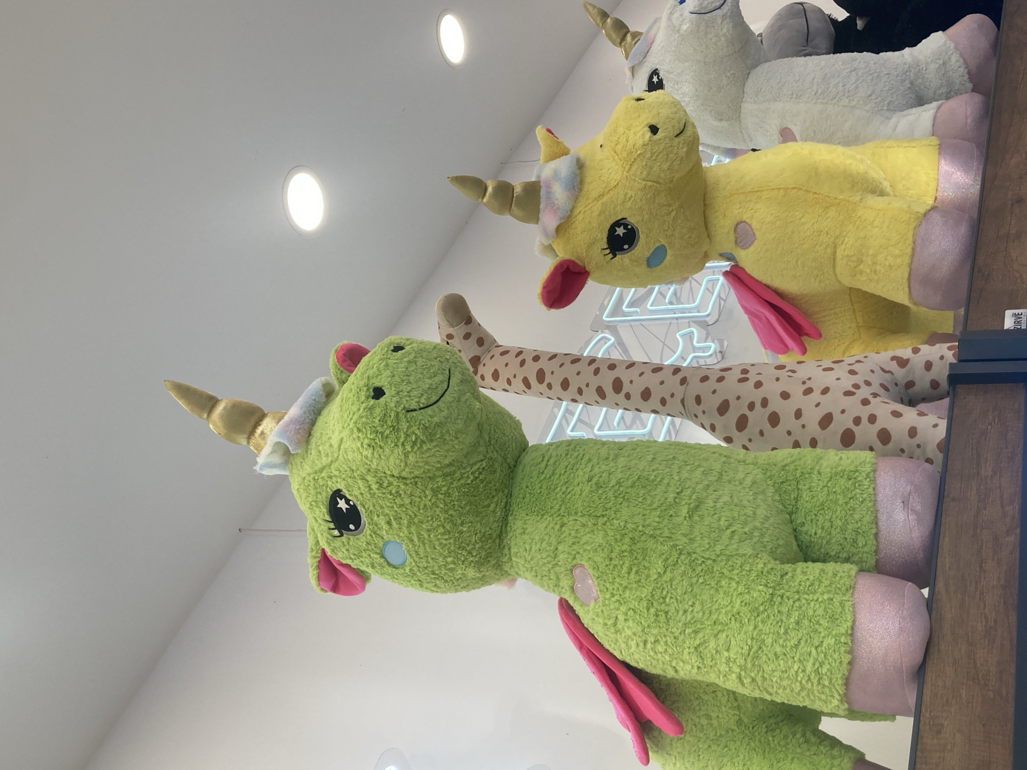 A low-angle, close-up shot shows a line of large, plush stuffed animals on a wooden shelf, illuminated by ceiling lights. In the foreground, a bright green unicorn with pink wings and hooves is positioned to the left. To its right, a spotted giraffe stands tall. Further to the right, a yellow unicorn with pink wings and hooves is visible, followed by a white unicorn with pink hooves. Behind the animals, a bright neon blue sign with partially visible letters is mounted on a white wall. The overall scene appears to be indoors, likely in a retail environment, possibly a toy store or gift shop. There are no people or actions depicted in the image. The location is not discernible from the image itself.