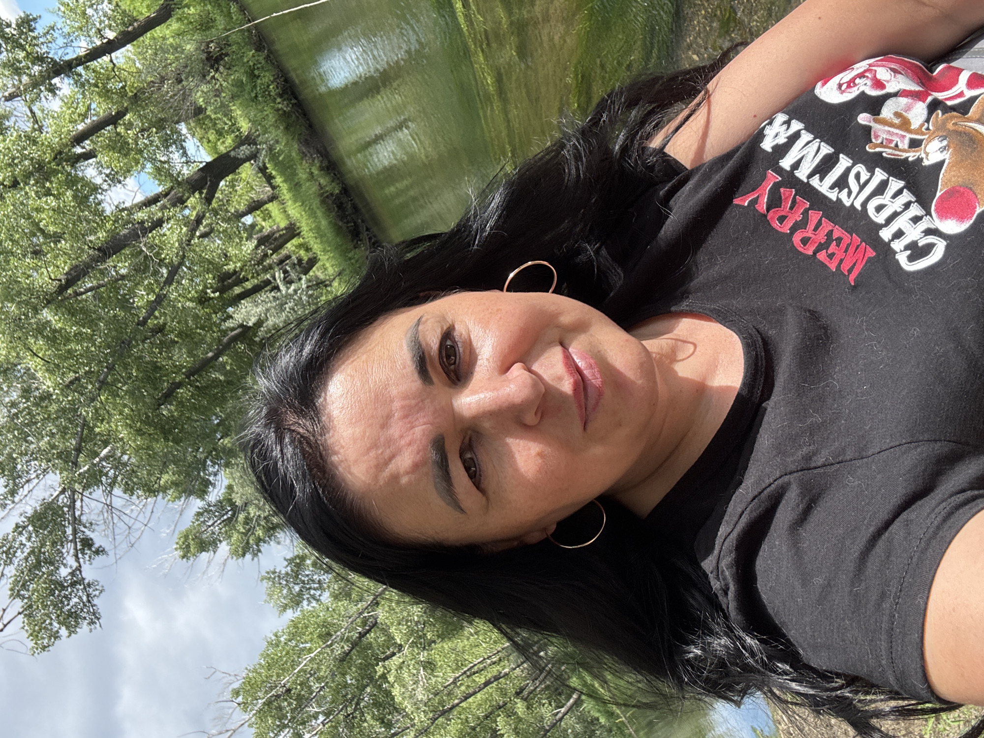 A woman with long, dark hair is taking a selfie outdoors. She is wearing a black t-shirt with a "Merry Christmas" graphic. Behind her, a body of water reflects the surrounding trees and sky. The location is identified as the city of Starokhalilovo, Russia.