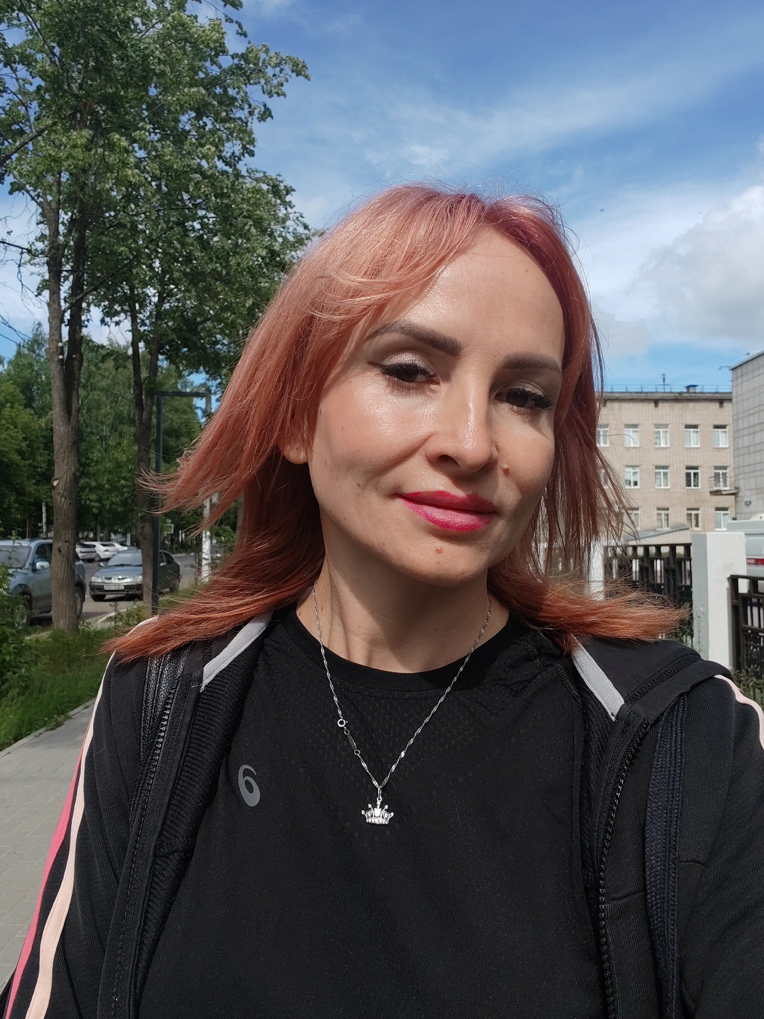A woman with reddish-pink hair is taking a selfie outdoors on a cloudy day. She is wearing a black shirt with a logo and a black jacket with pink and white stripes on the sleeves. A silver necklace with a crown pendant is visible. Behind her, there are trees, a sidewalk, parked cars, and buildings. The location is identified as Chaikovsky, Russia.