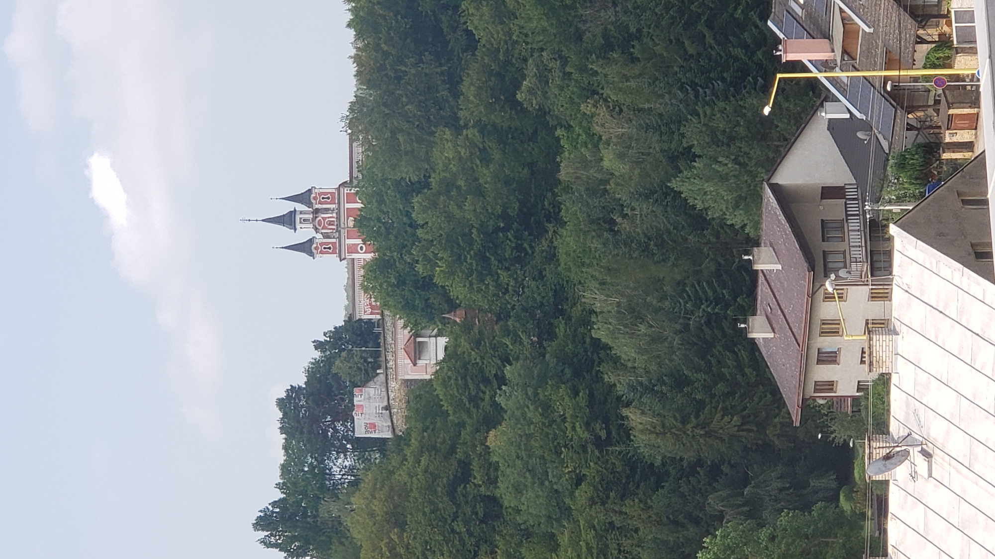 This is a vertical, medium-shot, outdoor, daytime photograph taken from a slightly elevated position overlooking a town nestled against a forested hillside. The sky is overcast with some wispy clouds.
In the upper portion of the frame, a red and white building with multiple spires, resembling a church or a historical monument, is visible atop a hill. The building has decorative architectural elements, including arched windows and ornate detailing on its facade. Below this structure, on the hillside, is a smaller building with a red-tiled roof and a small, open-air structure with a pitched roof supported by columns. Further down the slope, a wall constructed of stone blocks is visible, with some scaffolding and what appears to be building materials covered in white tarps.
The middle and lower portions of the image are dominated by a dense, dark green forest that covers the hillside. The trees are a mix of deciduous and coniferous varieties, creating a thick canopy.
In the foreground, at the bottom of the frame, several buildings with sloped, dark-colored roofs are visible. One prominent building has a light-colored facade, multiple windows, and chimneys. Another building to its right has a darker roof with solar panels. A bright yellow streetlamp with a circular red sign is positioned on the right side of the frame. Several satellite dishes are also visible on the rooftops.
While the specific location within the city of Prešov, Slovakia, is not explicitly identifiable from the image alone, the architectural style of the prominent building on the hill and the surrounding landscape suggest a European setting. It's plausible this is a view of a significant landmark or religious site overlooking the town, possibly with historical or cultural importance within Prešov.