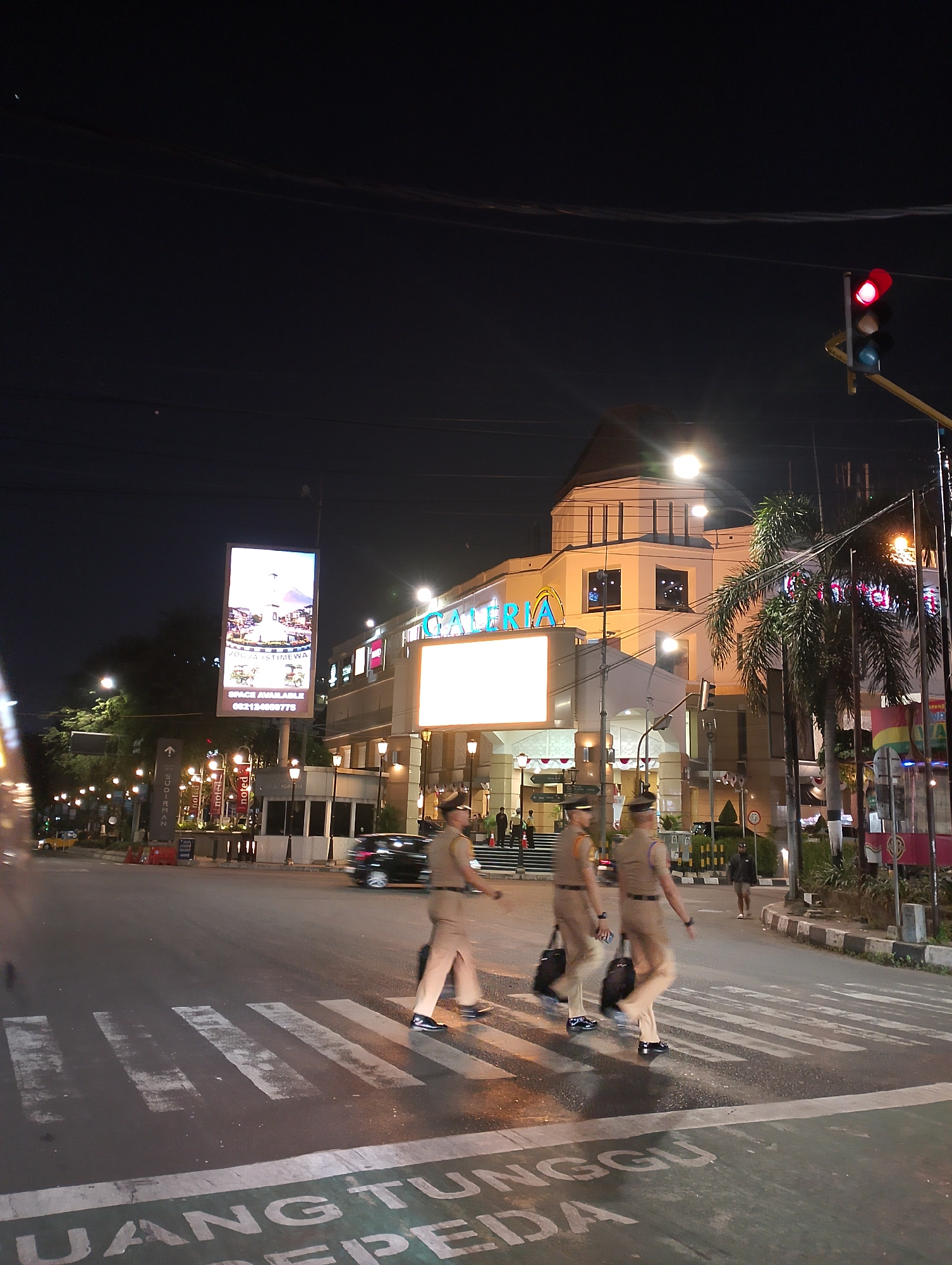 The image captures a nighttime scene at a pedestrian crossing in front of a building identified as "GALERIA" in Yogyakarta, Indonesia. Three individuals, appearing to be uniformed personnel, are crossing the street. They are wearing light-colored uniforms with hats and carrying dark bags. One person in the foreground is slightly blurred due to motion. A dark-colored car is visible in the background on the left side of the street. The building, "GALERIA," is illuminated with bright lights, and a large digital billboard is displayed on its facade. Traffic lights are present, with a red light illuminated. Palm trees and streetlights are also visible, contributing to the urban nighttime atmosphere. The road surface shows painted markings for the pedestrian crossing and text that appears to be in Indonesian.