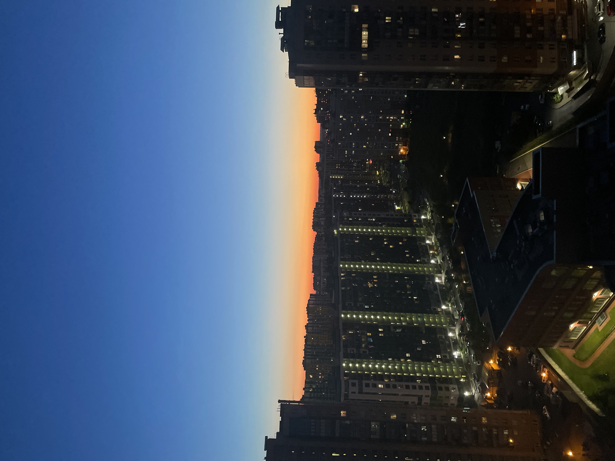 This is a high-angle, wide shot of a cityscape at dusk. The sky is a gradient of deep blue at the top, transitioning to orange and red near the horizon. In the foreground, several tall, modern apartment buildings are visible, with many windows illuminated. These buildings are densely packed together, suggesting an urban environment. Below and between the buildings, there are visible roads, parked cars, and some green spaces. The overall scene depicts a residential area in a city as evening sets in.