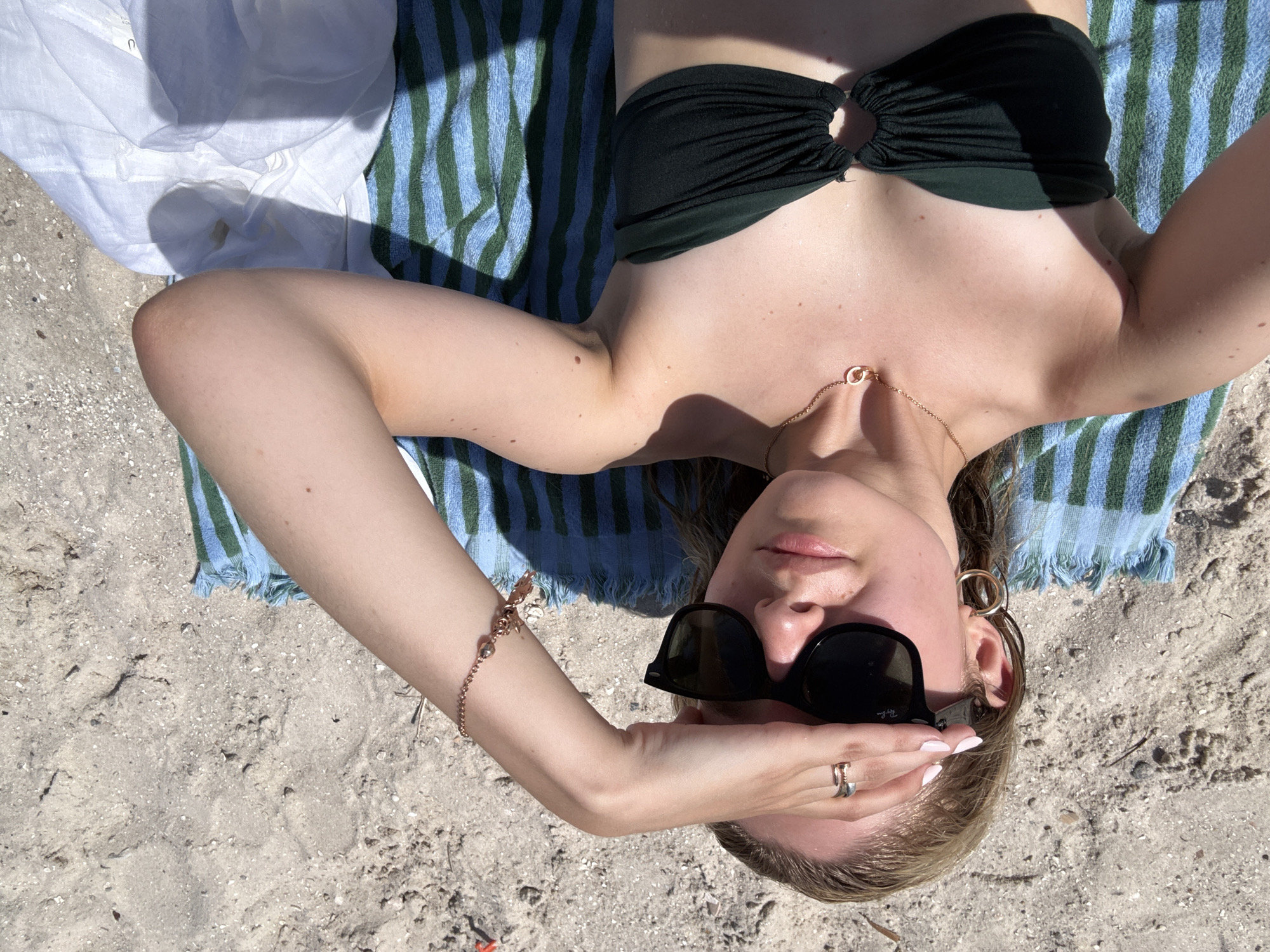 A woman lies on a striped towel on a sandy beach, with her right arm raised to shield her eyes from the sun. She is wearing sunglasses, a dark green bikini top, a gold necklace, hoop earrings, and rings on her fingers. A rose gold charm bracelet adorns her wrist. The scene is set on a beach in Miami, United States of America, under a clear sky.