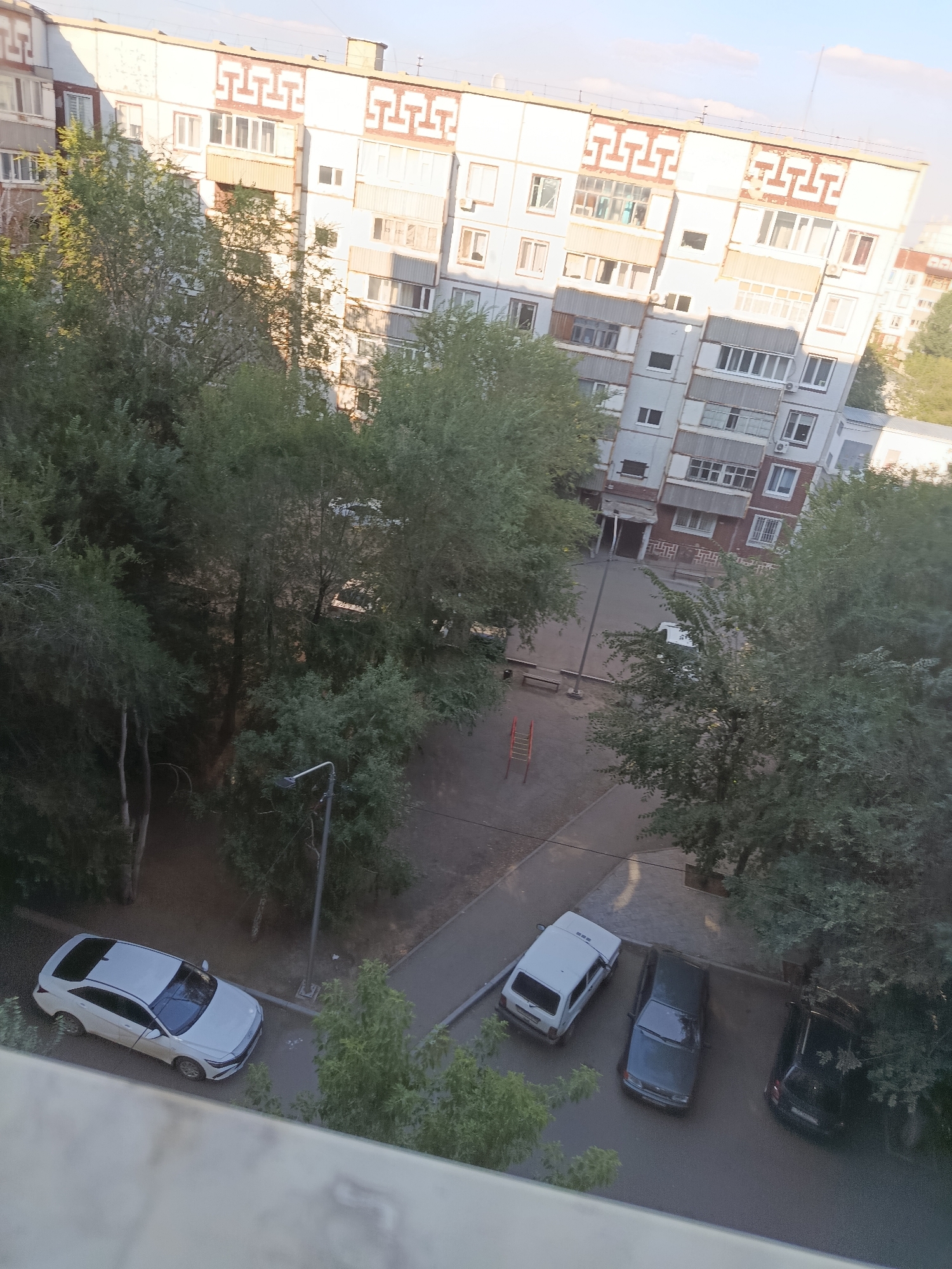 The image shows a residential courtyard in Ekibastuz, Kazakhstan. A multi-story apartment building with a light-colored facade and decorative patterns is visible on the right side of the frame. Several trees with green foliage are scattered throughout the courtyard. In the foreground, a white car is parked on the asphalt. To the right of the white car, there are two more cars, one dark gray and one black, parked in a row. A street lamp is also present. In the background, the courtyard features a small playground with a red slide. The scene appears to be a typical urban environment.