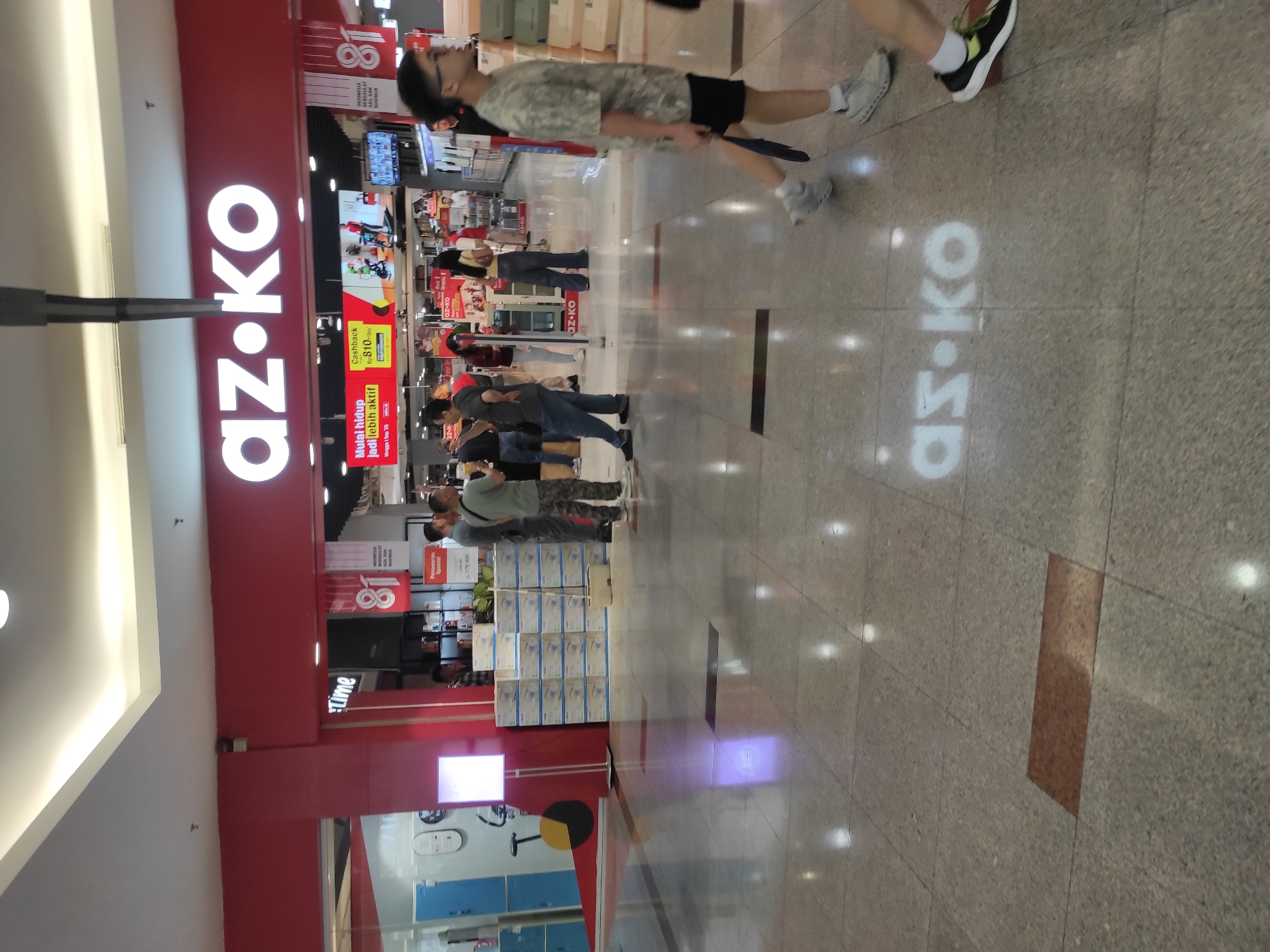 The image shows the entrance to an AO•KO store in the city of Surabaya, Indonesia. The store has a prominent red facade with the AO•KO logo in white lettering. Inside the store, there are several people browsing products. A variety of electronic items and appliances are displayed on shelves. The floor is tiled with a reflective surface, and the lighting in the store is bright. There are also promotional banners visible. The overall atmosphere appears to be that of a busy retail environment.
