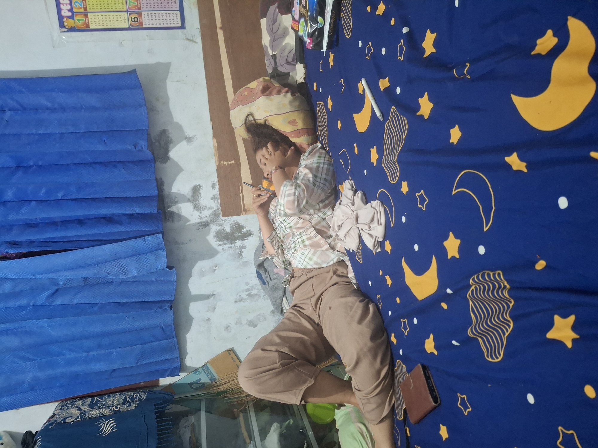 A woman is lying on a bed in what appears to be a bedroom. She is wearing tan pants and a plaid shirt. Her head is resting on a pillow, and she is holding a pen in her right hand, touching her face. The bed is covered with a blue sheet featuring yellow moons and stars. To the left of the bed, there is a wooden shelf unit with some items on it, and blue curtains hang above. On the wall to the right, there is a chart with numbers and text, possibly a learning aid. A wallet is placed on the bed near the woman's feet. The room has a neutral-colored wall and a wooden headboard.