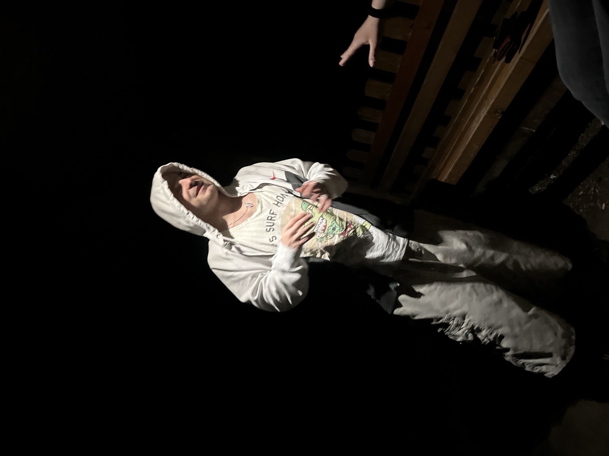 The dimly lit image captures a scene in Blagoevgrad, Bulgaria, likely at night. A young person is lying on their back, partially illuminated, with their head tilted back and mouth open, suggesting laughter or a relaxed state. They wear a white hooded sweatshirt with the hood up, light-colored pants, and a white t-shirt. Visible text on the t-shirt reads "S SURF HOM". A silver necklace is also visible around their neck. Their hands are resting on their chest/stomach area.
In the upper right portion of the frame, another person's arm and hand are visible, reaching downwards towards the individual on the ground, indicating an interaction. Part of their dark-colored pants is also visible. Behind the reaching hand, a wooden structure composed of dark, horizontal slats can be seen, possibly part of a bench, bed frame, or railing. A small, dark object with red elements is located near this wooden structure. The rest of the scene is consumed by deep shadow and darkness.