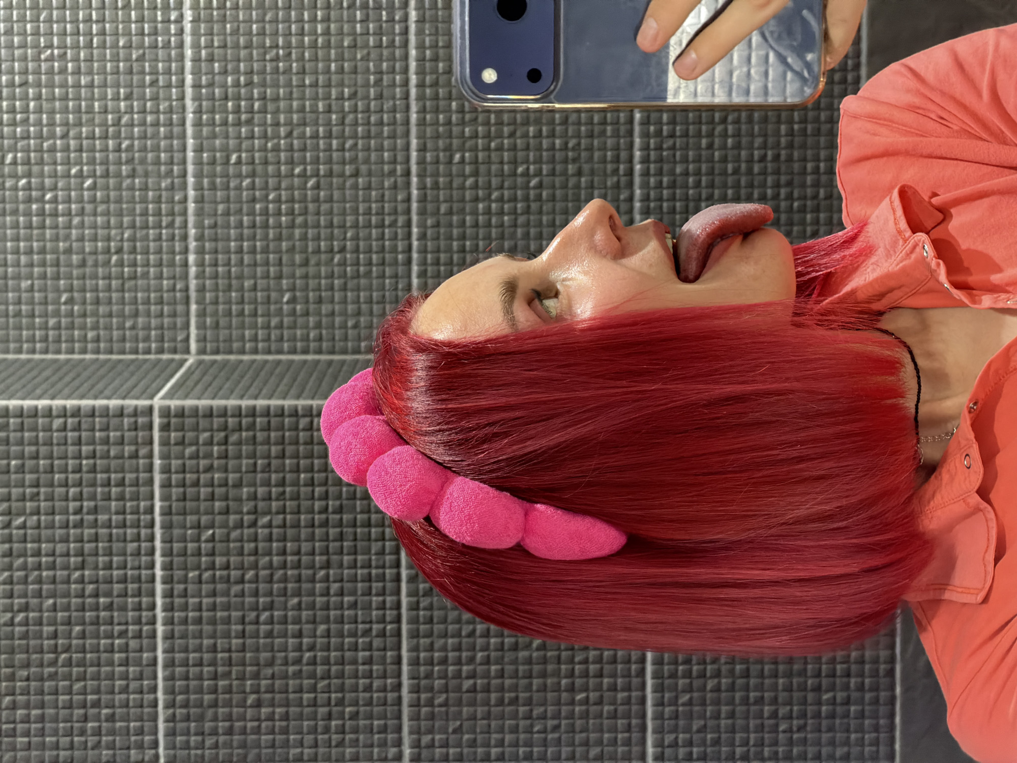The image depicts a person, reportedly located in the city of Chaikovsky, Russia, captured in profile, facing towards the right. The individual has vivid magenta-pink, bob-length hair and wears a bright pink headband composed of multiple large, plush, spherical elements. Their mouth is open, and a long tongue is extended downwards and slightly outwards. They are dressed in a coral-colored collared shirt with visible snap fasteners and a thin black choker necklace. On the right side of the frame, a dark blue smartphone is held up by a partially visible hand, with its camera module clearly discernible, reflecting part of the scene. The background consists of a wall covered in small, dark grey, square-textured tiles, bisected by a prominent horizontal grout line. Diagonal lines are visible on the far right, indicating an architectural feature or different surface treatment.