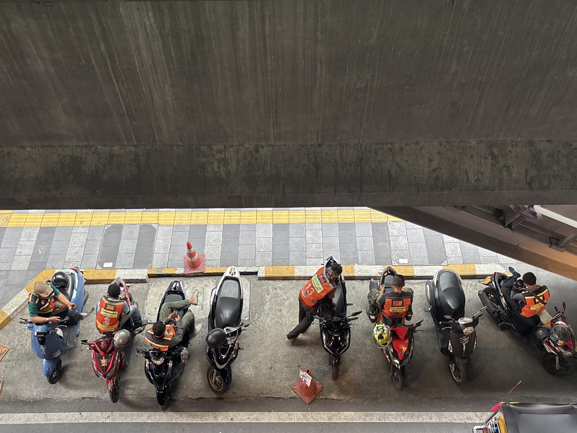 An overhead view captures a line of motorcycles parked along a sidewalk adjacent to a textured concrete wall. The scene is in Bangkok, Thailand. Several individuals, wearing vests, are seated on or near their motorcycles, some appearing to be using mobile phones. The sidewalk features a tactile paving strip with raised yellow bumps. Traffic cones are visible on the ground near the motorcycles. The overall impression is one of a temporary pause in activity for the motorcyclists.