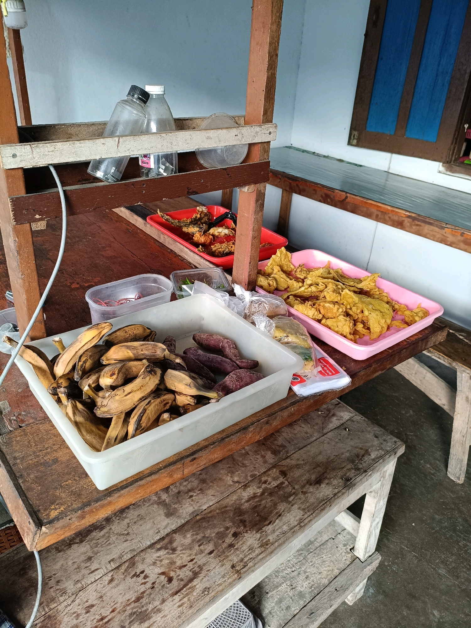 This is a forensic, matter-of-fact description of a food stall in Maguwoharjo, Indonesia. The stall is constructed from rough, weathered wood and is set up outdoors, likely on a street or sidewalk. The scene captures a selection of food items displayed for sale.
In the foreground, a white plastic rectangular container holds a collection of ripe bananas, some appearing overripe with brown spots. Adjacent to the bananas, also within the same container, are several dark purple-skinned sweet potatoes. To the left of this container, a smaller clear plastic container holds a bundle of red rubber bands. Further to the left, a white electrical cord snakes downwards from a light fixture.
Behind the bananas and sweet potatoes, a red plastic tray contains what appears to be fried fish pieces. To the right of this tray, a pink plastic tray is filled with a generous portion of golden-brown fried food, possibly tempura or fried snacks. Some of these fried items appear to be made with vegetables. A clear plastic bag, partially filled with the same fried items, is placed between the white and pink trays.
On a shelf above the food, two clear plastic bottles are visible, one with a black cap and the other with a white cap. These appear to be repurposed water bottles.
The stall itself is supported by wooden posts and planks. In the background, a wall painted white and a wooden window frame with blue slats are visible. The ground beneath the stall is concrete. The overall impression is of a simple, utilitarian food stall operating in a public area. No people are visible in the image.