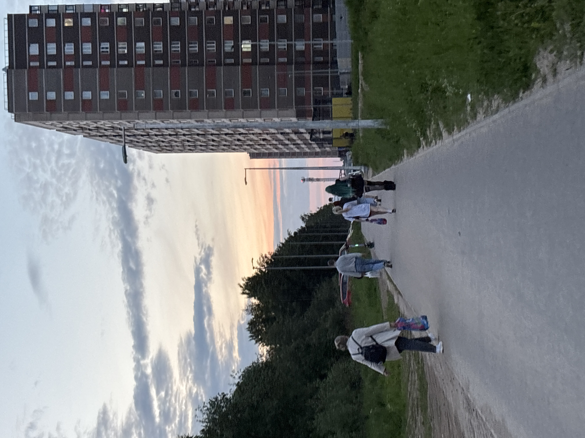 This image captures a late evening scene in Sinyavino, Russia, along a wide paved pathway. A group of approximately five young individuals is visible, some carrying or pushing longboards or skateboards, as they walk away from the viewer along the path.
To the right of the path, a gentle grassy slope extends upwards. To the left, a dense line of trees borders the pathway, beyond which stands a large, multi-story residential building with a repetitive facade of numerous windows, suggesting a typical planned urban or suburban environment. The architecture hints at a functional, possibly Brutalist, design common in post-Soviet housing.
The time of day is clearly sunset or late dusk, evidenced by the warm hues of orange, pink, and pale blue painting the horizon above the distant trees and behind the apartment building. The sky further overhead is lighter, with scattered, elongated clouds. The overall mood appears relaxed and casual, with no overt emotional expressions discernible from the distance. Several street lights are visible in the midground, further indicating the transition from day to night. There is no visible text in the image.