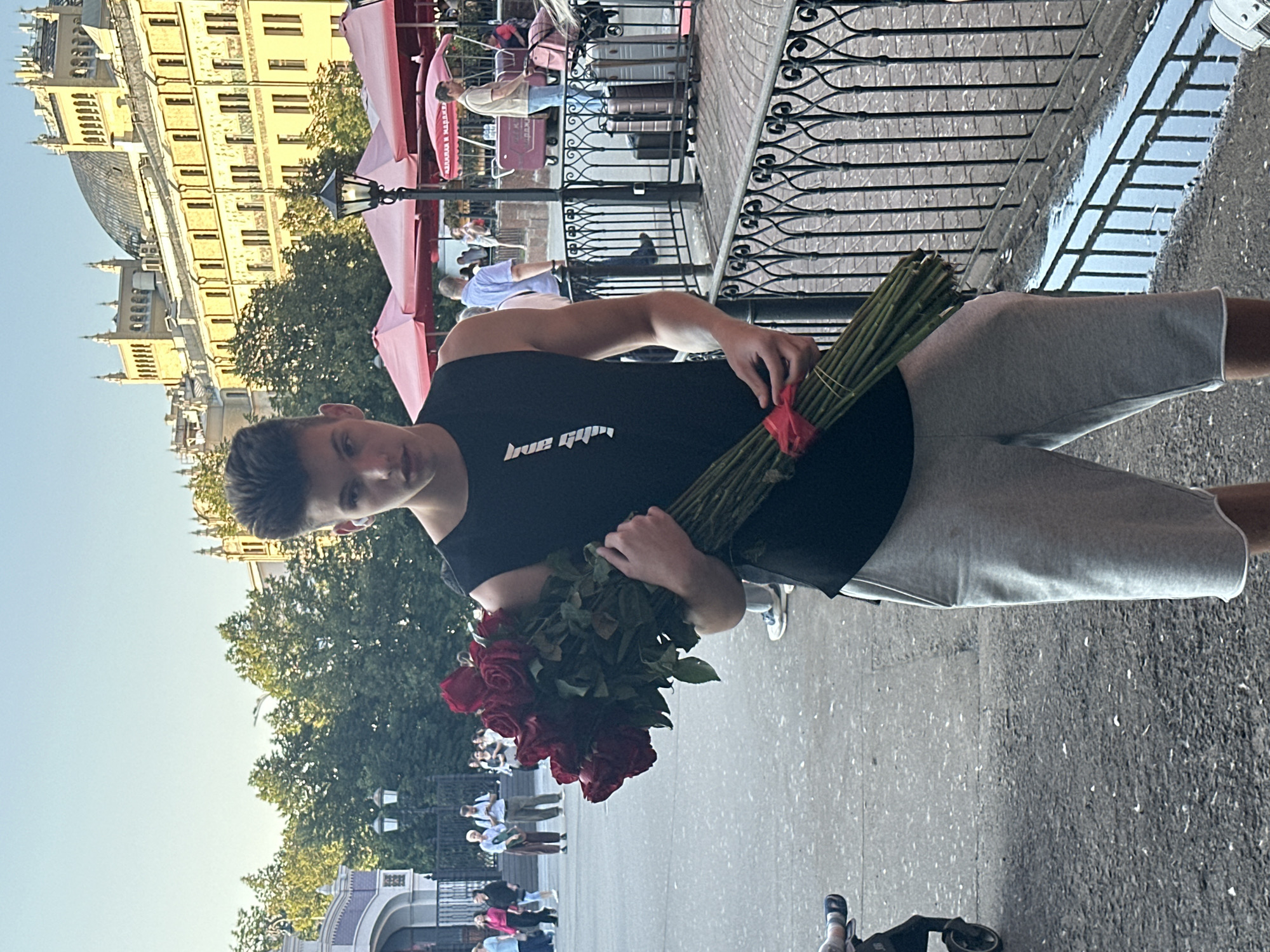 A young man with short, dark hair and a black sleeveless shirt stands in the foreground, holding a large bouquet of red roses. He is wearing light gray shorts. The background shows a bustling city street with pedestrians, a building with ornate architecture, and outdoor seating with red umbrellas. The scene appears to be in Moscow, Russia, possibly near a cafe or marketplace, with a street lamp and a decorative metal fence visible. The pavement is wet in places, suggesting recent rain.