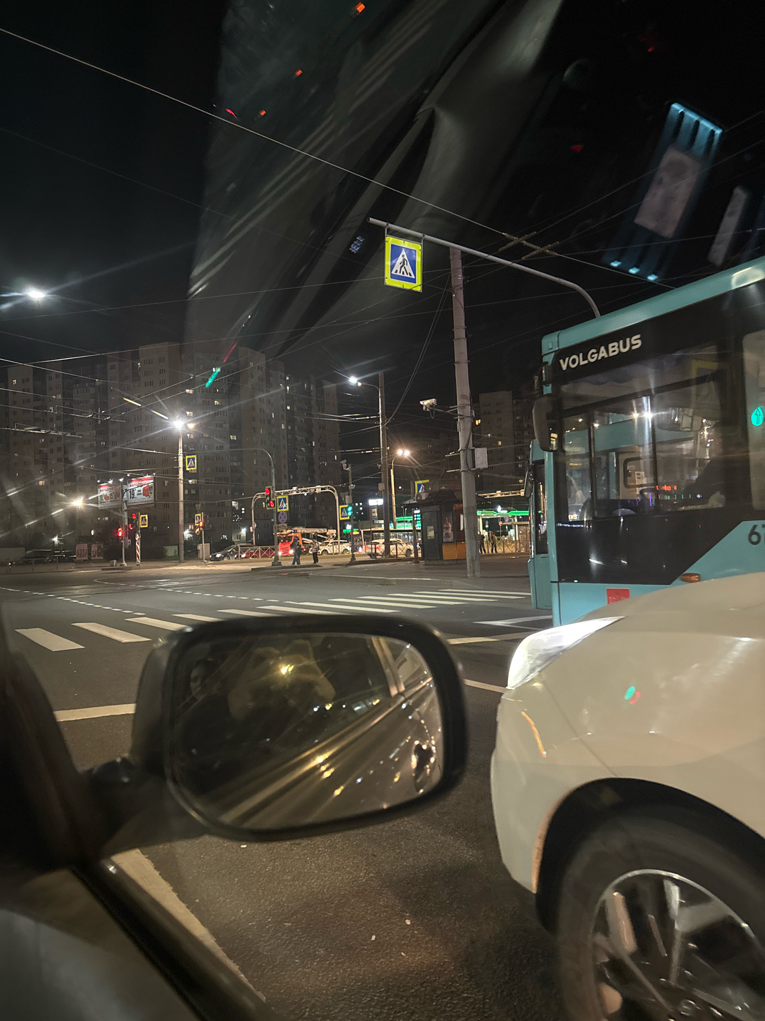 The image captures a street scene at night in Saint Petersburg, Russia. A turquoise public bus with "VOLGABUS" written on its side is prominently visible on the right side of the frame. The bus appears to be stopped, possibly at an intersection. In the foreground, the hood and wheel of a white car are visible, suggesting the photograph was taken from inside another vehicle. A side mirror of the car reflects the surrounding environment, including lights and what appear to be buildings.
The background shows a multi-lane intersection with traffic lights, pedestrian crossings, and overhead wires. Tall residential buildings with numerous windows are illuminated in the distance. The presence of traffic lights, pedestrian signs, and streetlights indicates an urban setting. The overall atmosphere is that of a busy city street at night, with the glow of artificial lights illuminating the scene.