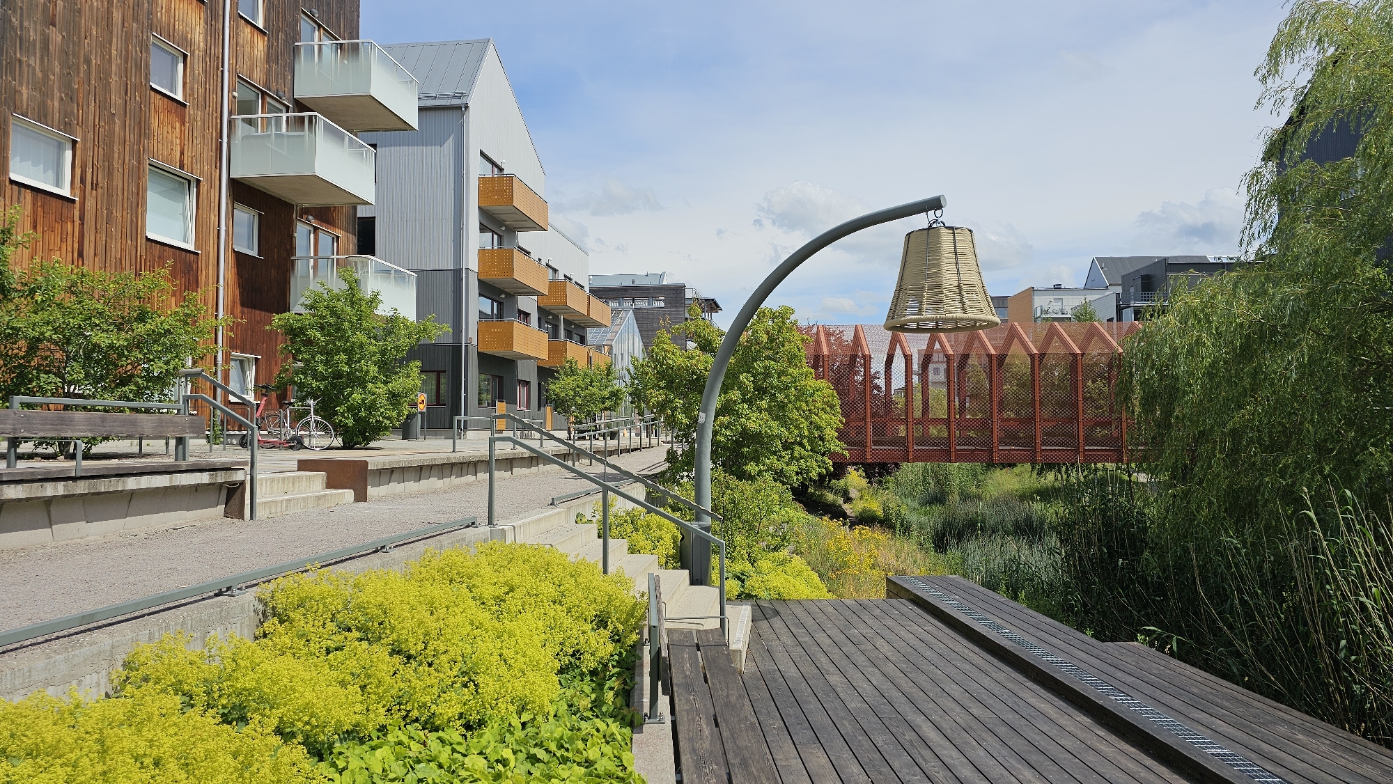 The image depicts a modern residential area adjacent to a canal or waterway in Linköping, Sweden. On the left, a multi-story building with a wooden facade and balconies with white railings is visible. Next to it, a building with a grey corrugated metal facade and balconies with orange accents stands. A paved walkway with a railing runs alongside the buildings, with a wooden bench to the left and a bicycle resting against some greenery.
In the center and right of the image, a distinctive red pedestrian bridge with an intricate lattice pattern spans the waterway. The bridge is surrounded by lush green trees and vegetation, including tall reeds near the water's edge. A large, woven lampshade hangs from a curved metal pole, positioned in the foreground overlooking the bridge and waterway. A wooden deck with a grate runs along the right side of the foreground. The sky above is partly cloudy.