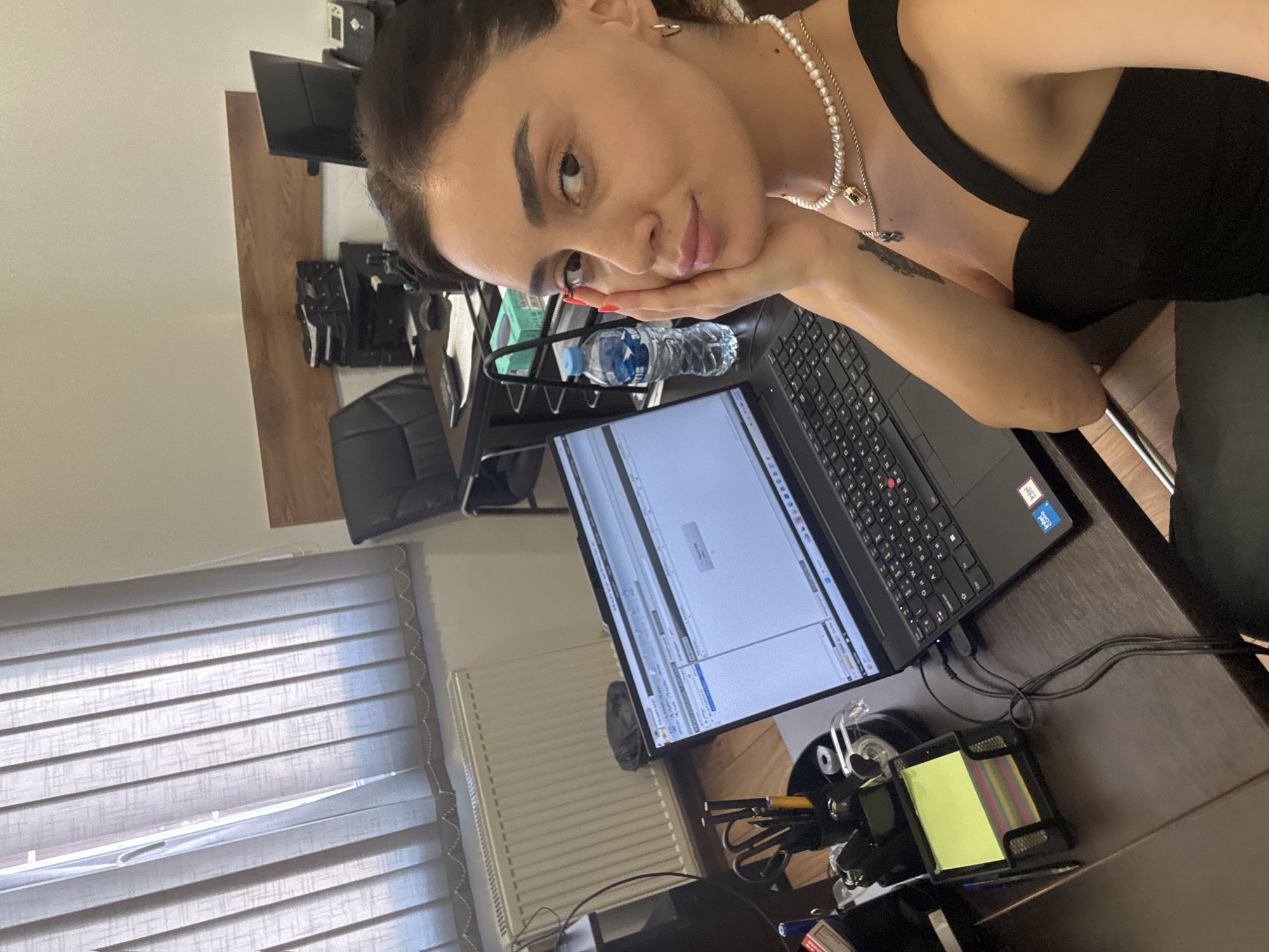 A woman is seated at a desk in an office. She is facing the camera, with her right hand resting on her cheek. She is wearing a black top and a pearl necklace. A laptop is open in front of her, displaying a spreadsheet. To her left, there is a desk organizer with pens and scissors, a stack of colorful sticky notes, and a pen. A bottle of water is on the desk to her right. In the background, there are vertical blinds covering a window, a radiator, and office furniture, including a black office chair and a desk with computer monitors. The scene appears to be an office environment in Tbilisi, Georgia, though no specific landmarks are visible to confirm this location.
