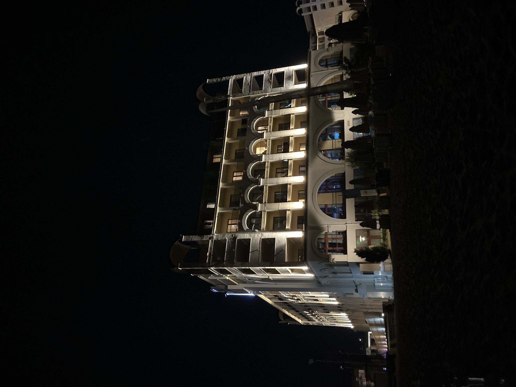The image depicts a multi-story building, likely a hotel, illuminated at night on a beach in Obzor, Bulgaria. The building features a facade with numerous arched windows and balconies, accented by bright exterior lighting. In front of the building, a beach area is visible with scattered beach chairs and umbrellas, some of which are closed. The sky is dark, indicating nighttime, and the overall scene is dimly lit, with the building being the primary source of light. There are no people visible in the image.