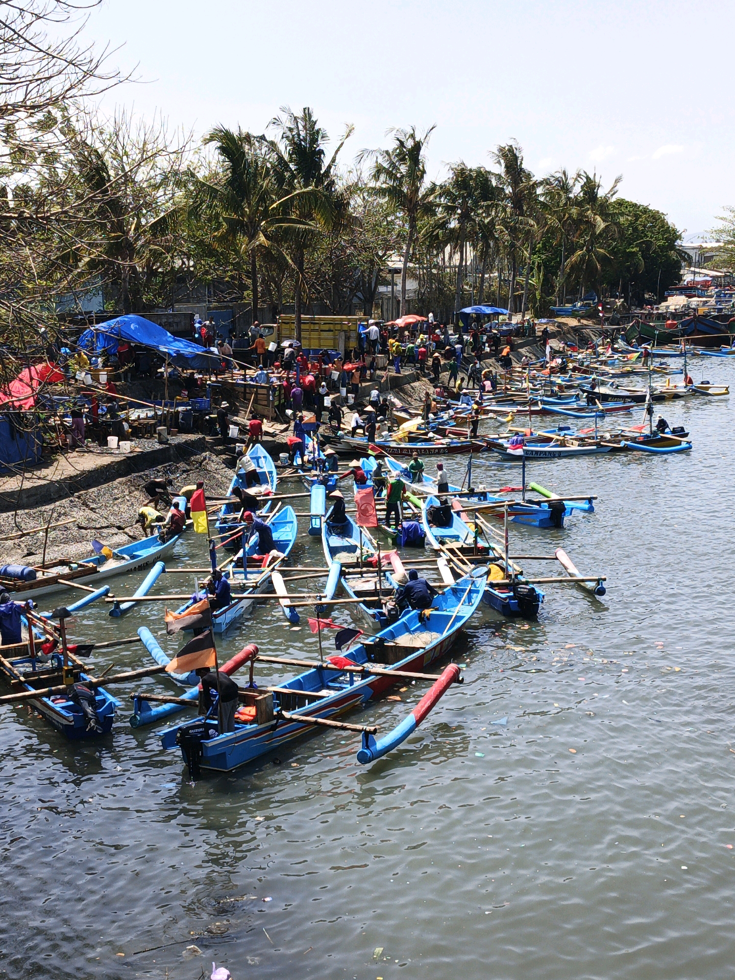 The image depicts a bustling scene at a waterfront, likely a fishing harbor or market, in Kelurahan Tegalkamulyan, Cilacap, Indonesia. Numerous small, brightly colored fishing boats, primarily blue with red and yellow accents, are docked side-by-side in the calm water. Many boats are equipped with outriggers and some have outboard motors. People, dressed in casual attire, are actively engaged in various activities. Some individuals are in the boats, possibly preparing them for departure, mending nets, or unloading catch. Others are on the shore, which is lined with makeshift stalls covered by blue and red tarpaulins. These stalls appear to be selling goods or processing fish. Palm trees and other vegetation are visible along the shoreline, with a few bare trees in the foreground. The sky is clear and bright, suggesting a sunny day. The presence of a yellow truck on the shore indicates the transport of goods. The water contains some scattered debris, typical of a busy port environment.